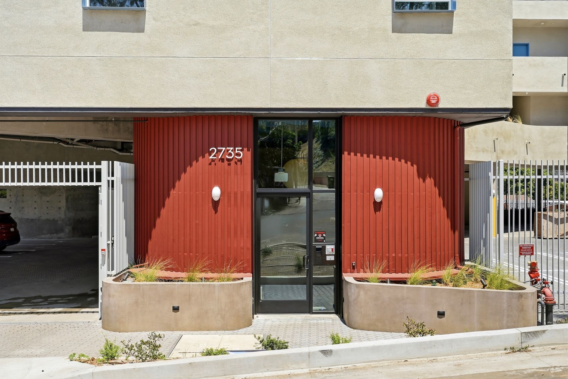 Building entrance with red paneling, glass door, and concrete planters by a sidewalk