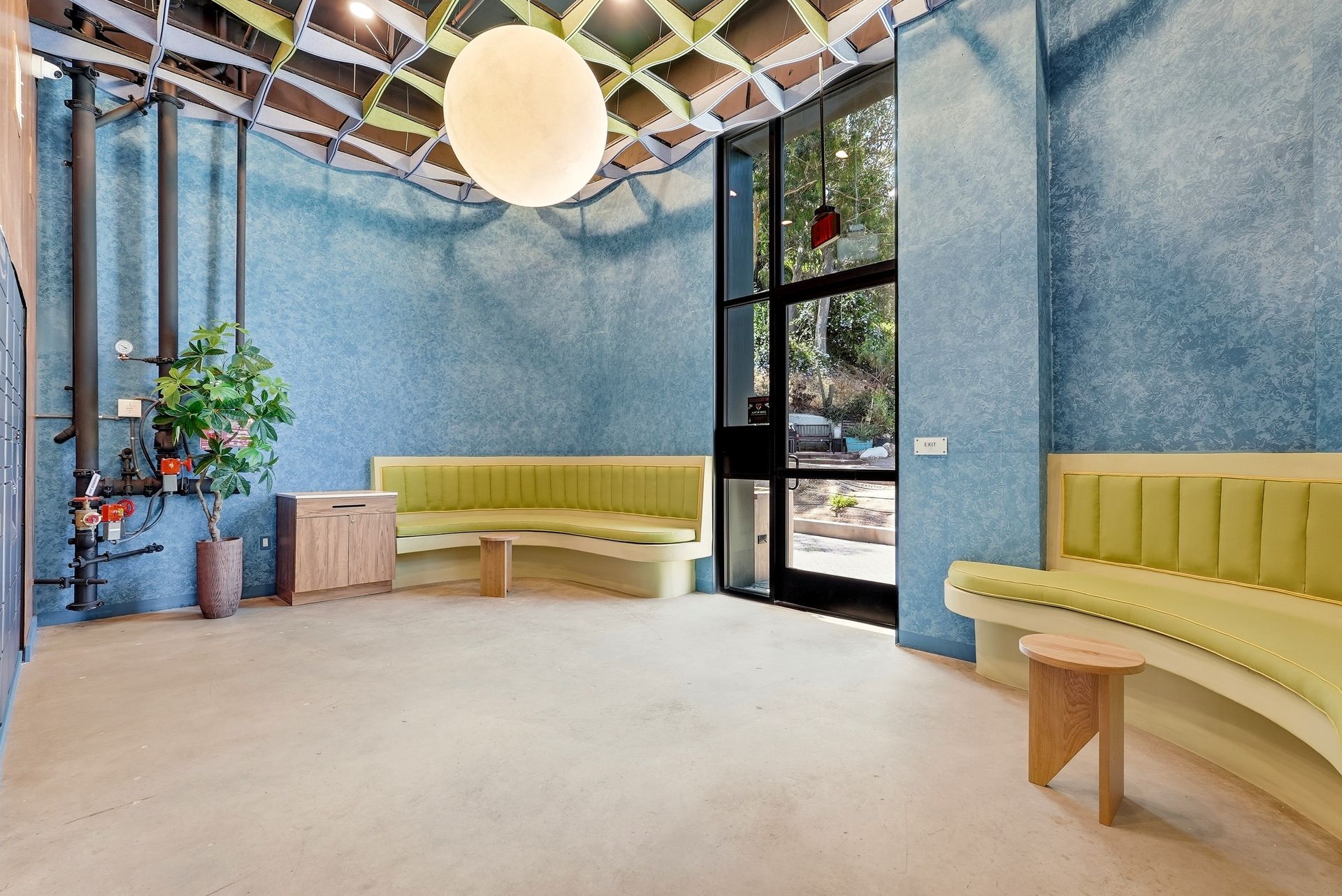 Modern lounge with blue walls, yellow curved benches, large windows, plants, and a hanging globe light.