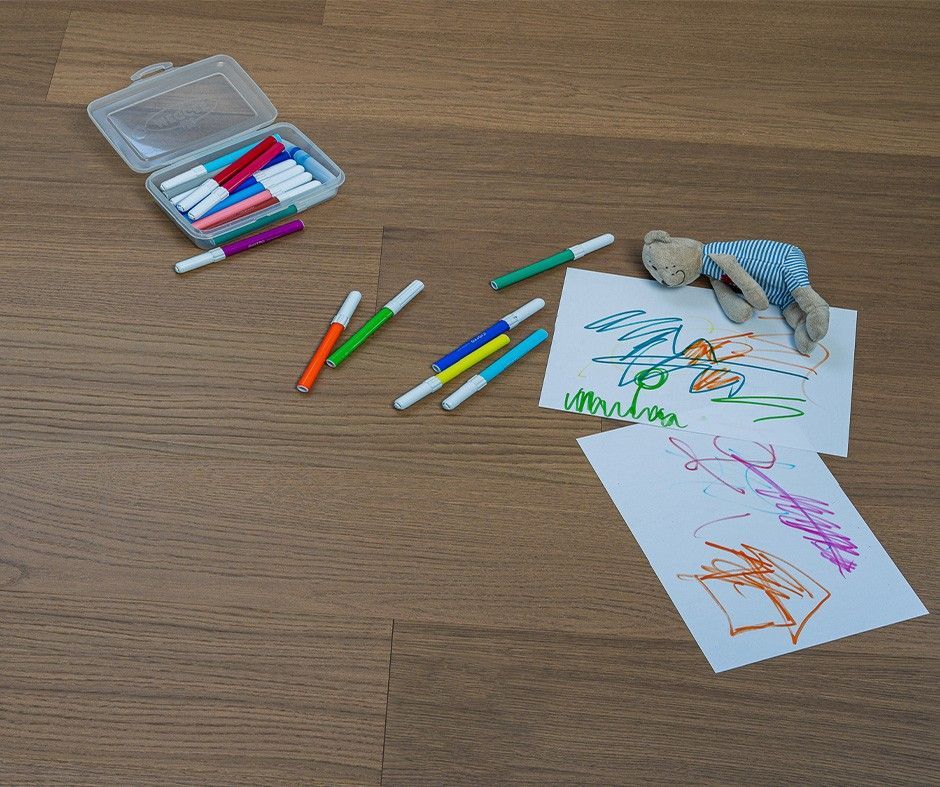 Pennarelli, disegni e un peluche su un pavimento di legno. Alcuni pennarelli sono sparsi, altri in una custodia aperta. I disegni sono colorati.
