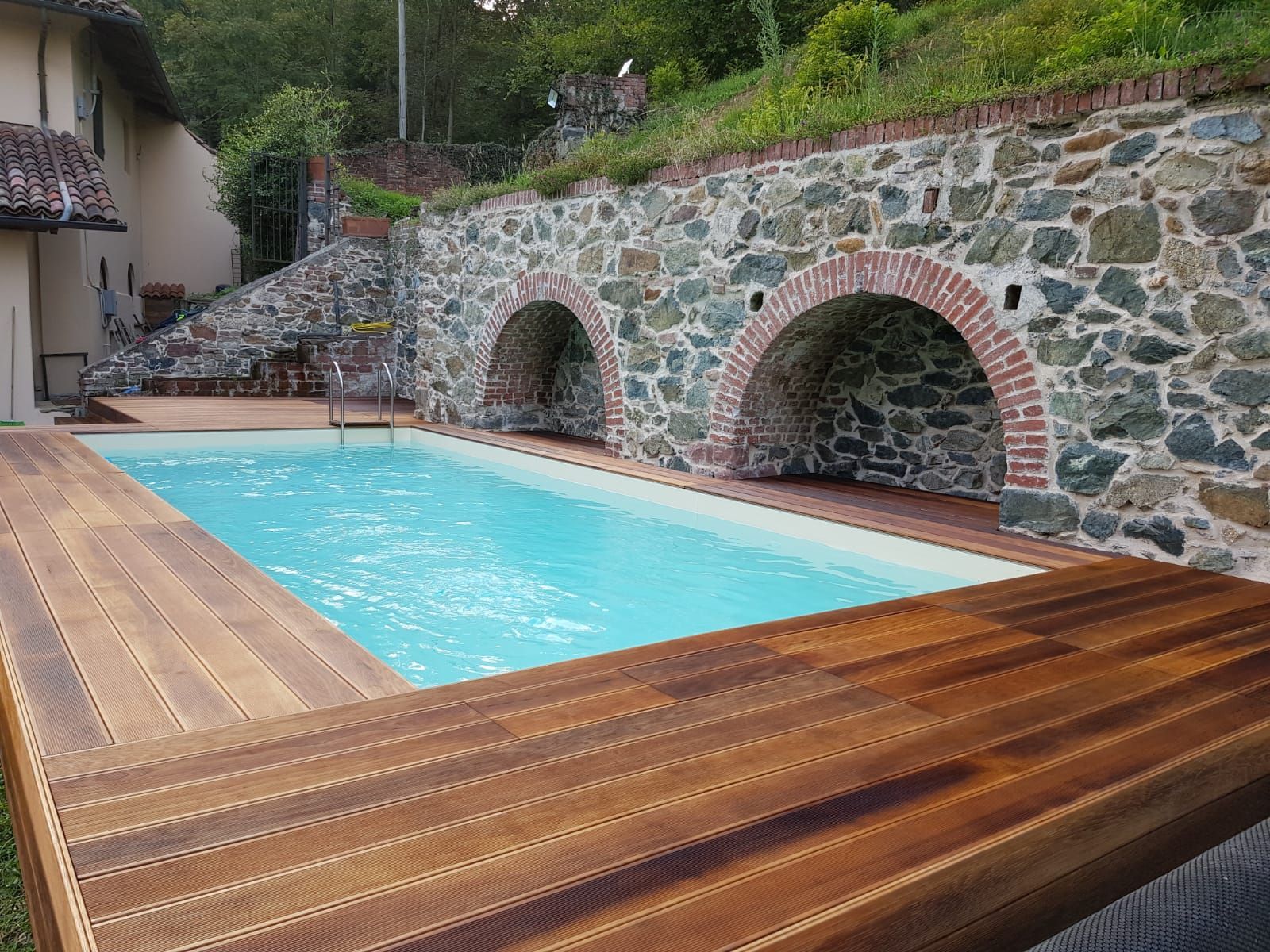 Piscina a sfioro con vista sulla città al tramonto. Acqua azzurra, terrazza in legno e architettura moderna.
