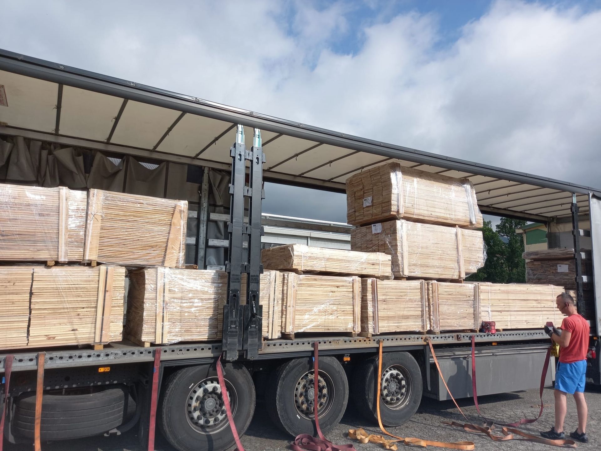 Un autoarticolato carico di pile di assi di legno, fissate con cinghie, sotto un cielo azzurro. Una persona con una maglietta rossa è in piedi lì vicino.