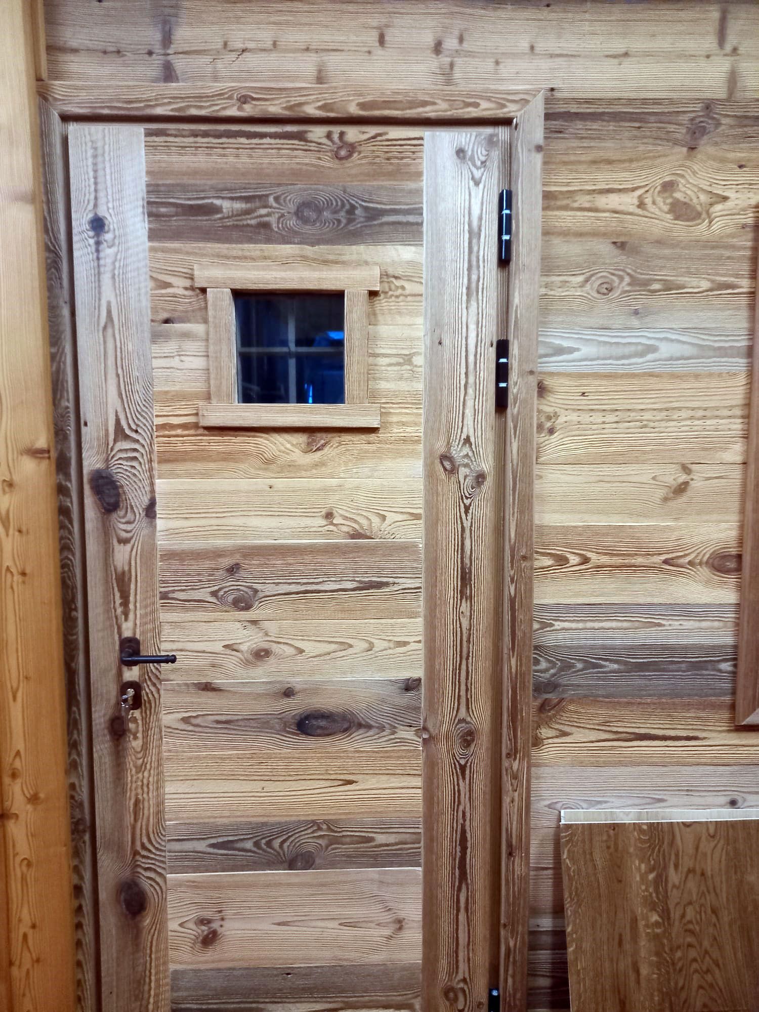 Porta in legno con una piccola finestra quadrata. Venature del legno marrone scuro e beige, con ferramenta nera.