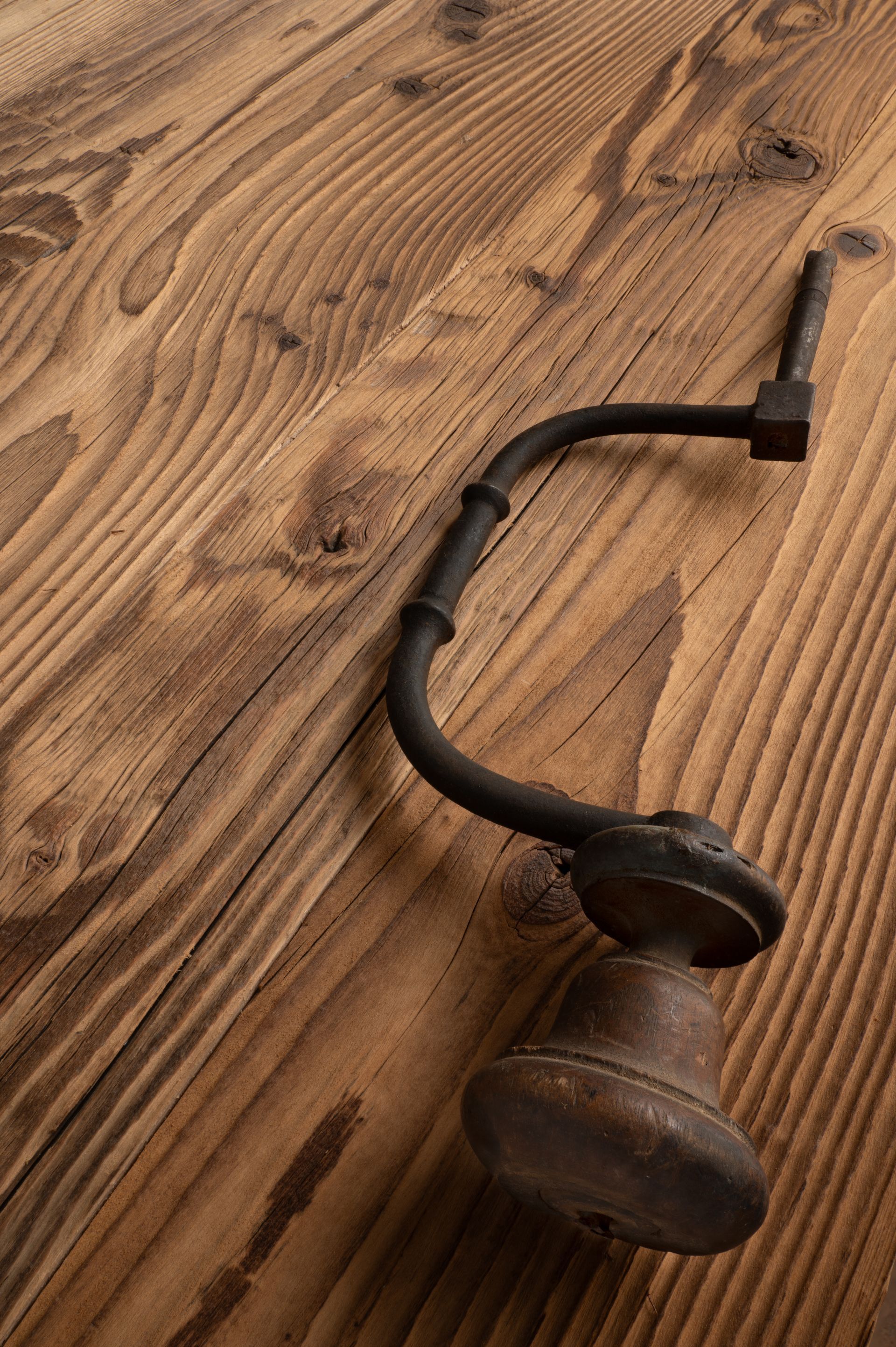 Un trapano a mano antico e arrugginito appoggiato su una superficie di legno con venature visibili.