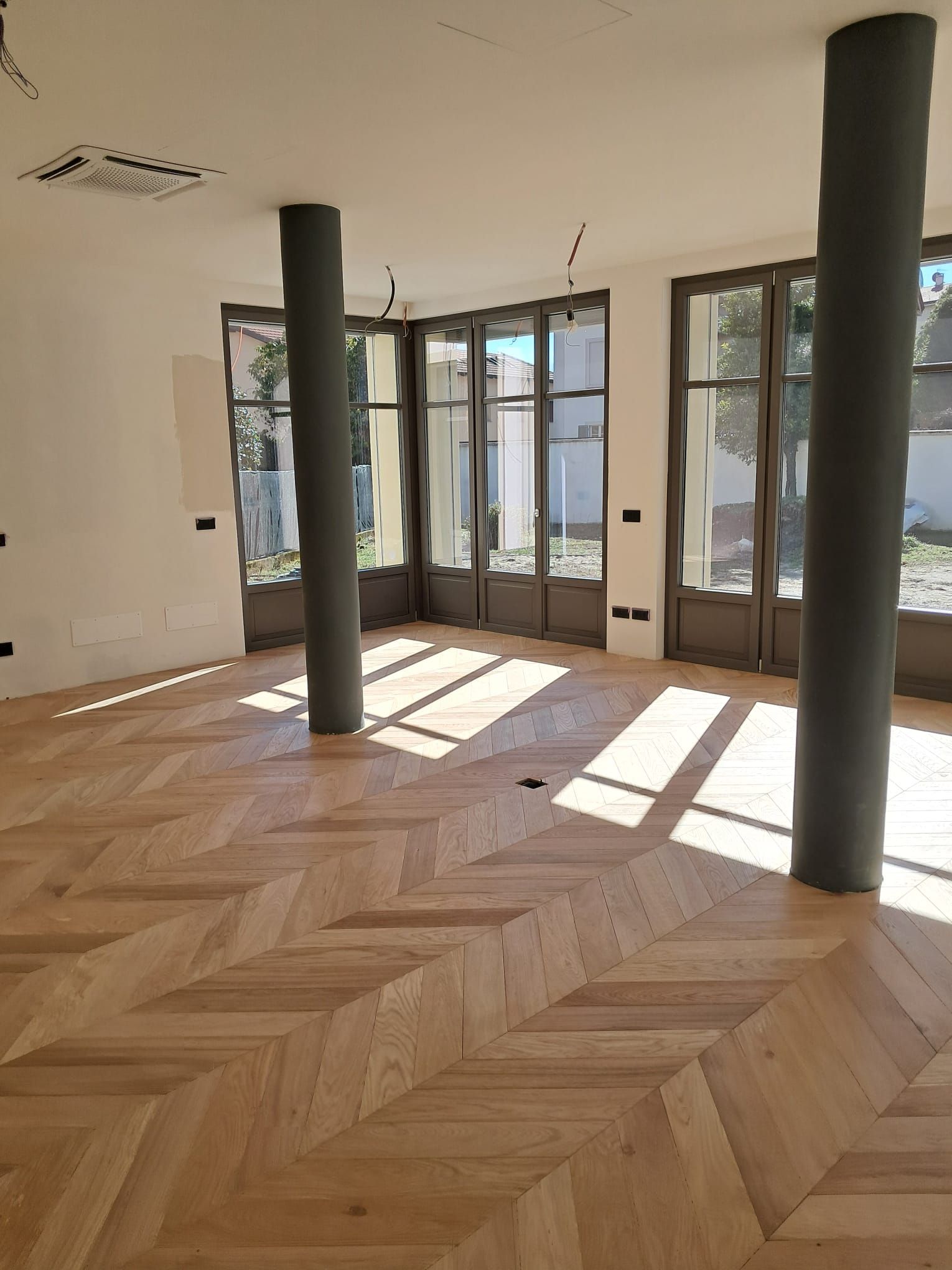 Stanza vuota con pavimento in legno chiaro a spina di pesce, due colonne grigie e alte porte a vetri. 