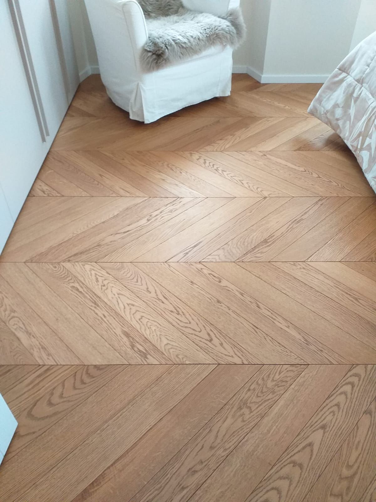 Pavimento in legno a spina di pesce in una stanza, con una poltrona bianca e parte di un letto visibili.