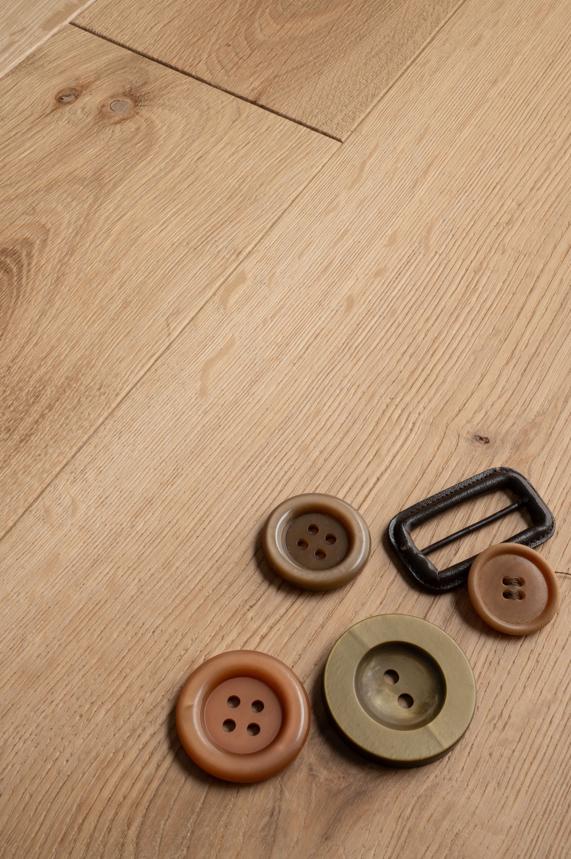 Quattro bottoni marroni e una fibbia rettangolare nera su una superficie con venature di legno chiaro.