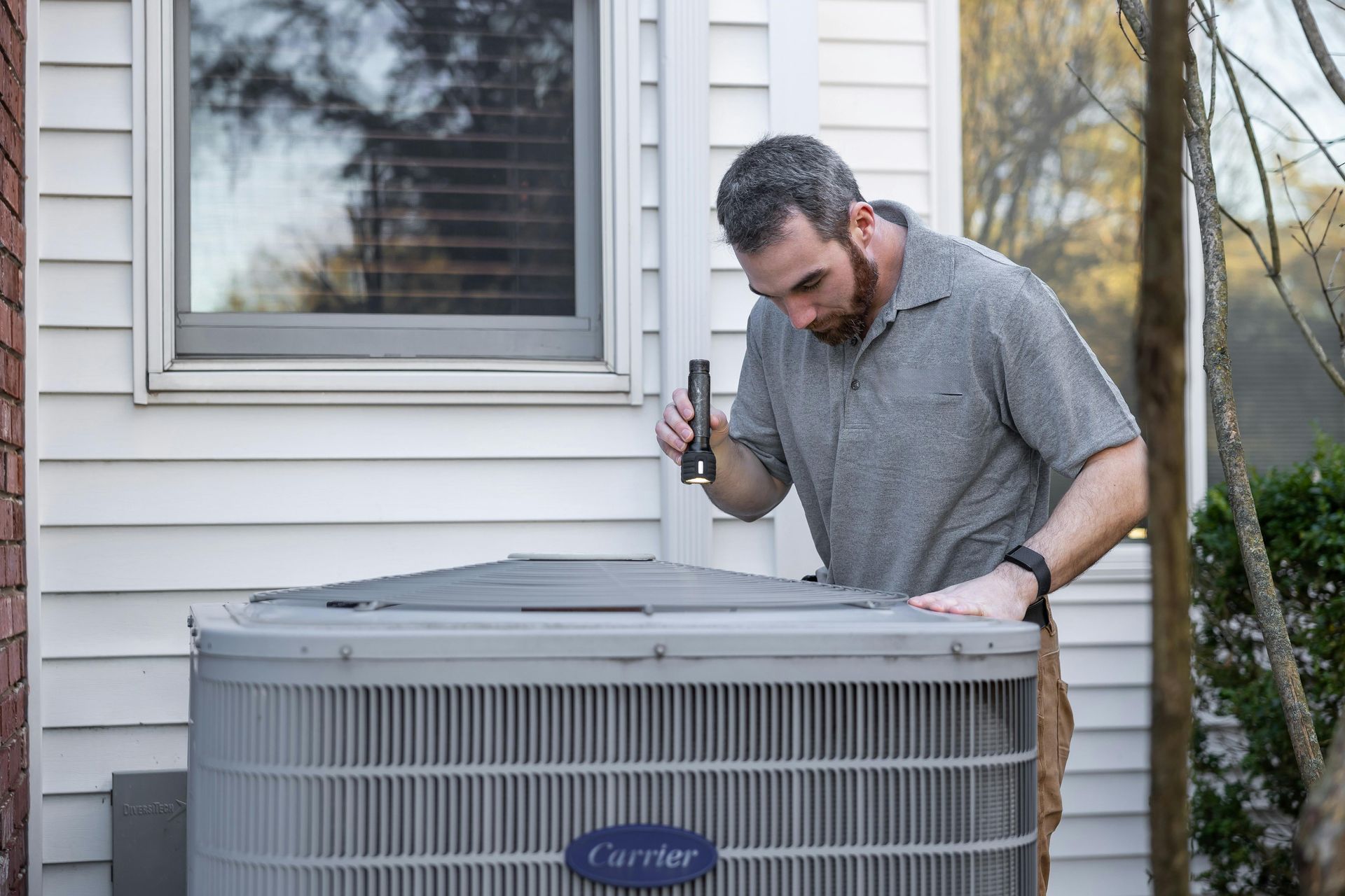 HVAC technician inspecting an air conditioning unit with a flashlight outside a house.