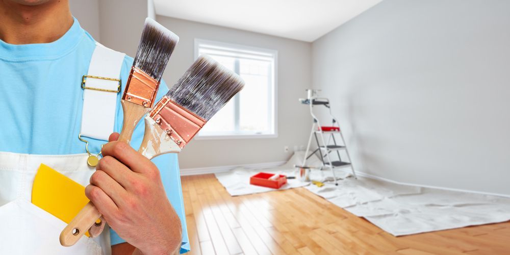 A man is holding two paint brushes in front of a room that is being painted.