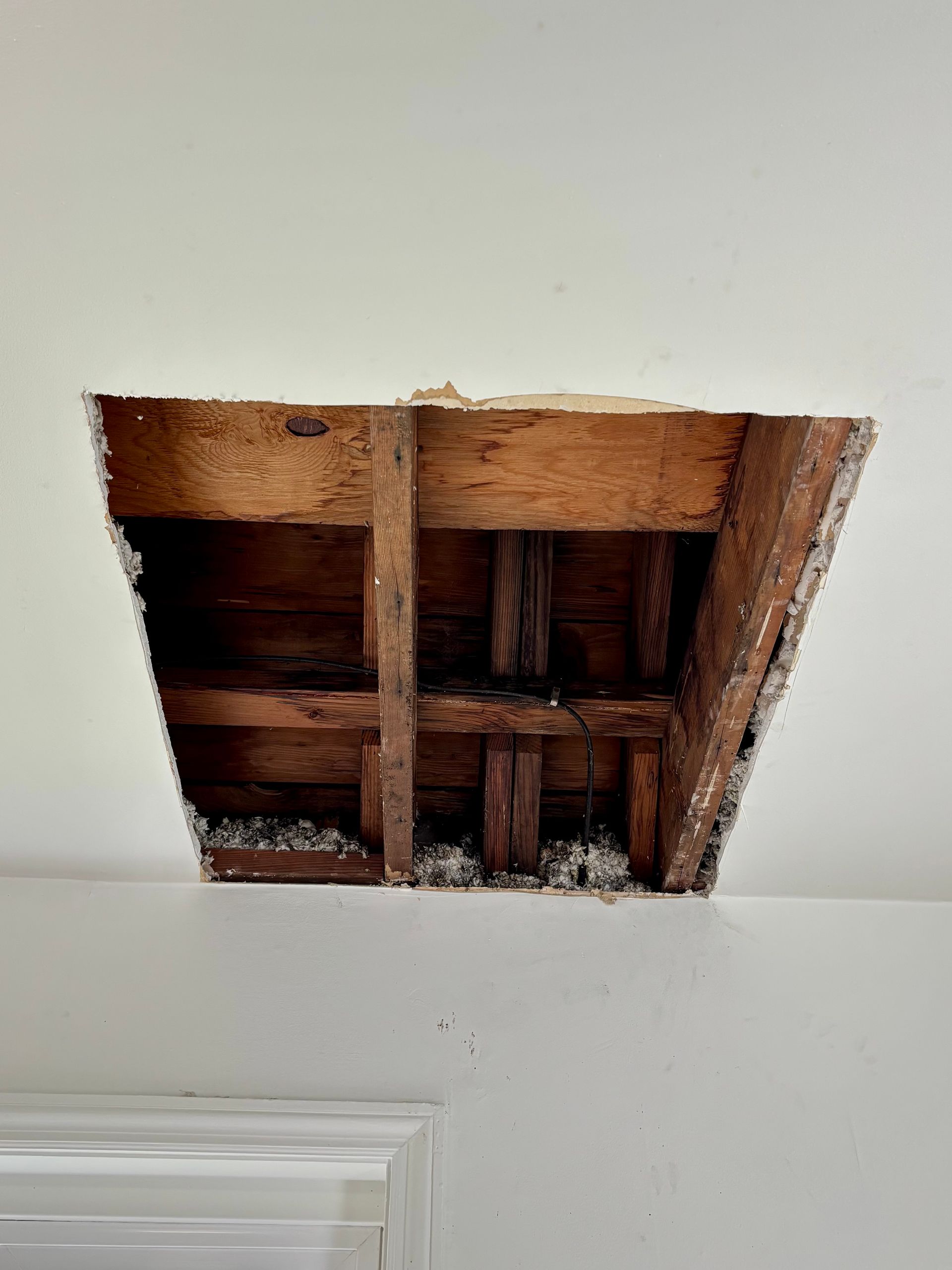 Hole in white ceiling exposing wooden beams and support structure. White paint flakes visible.