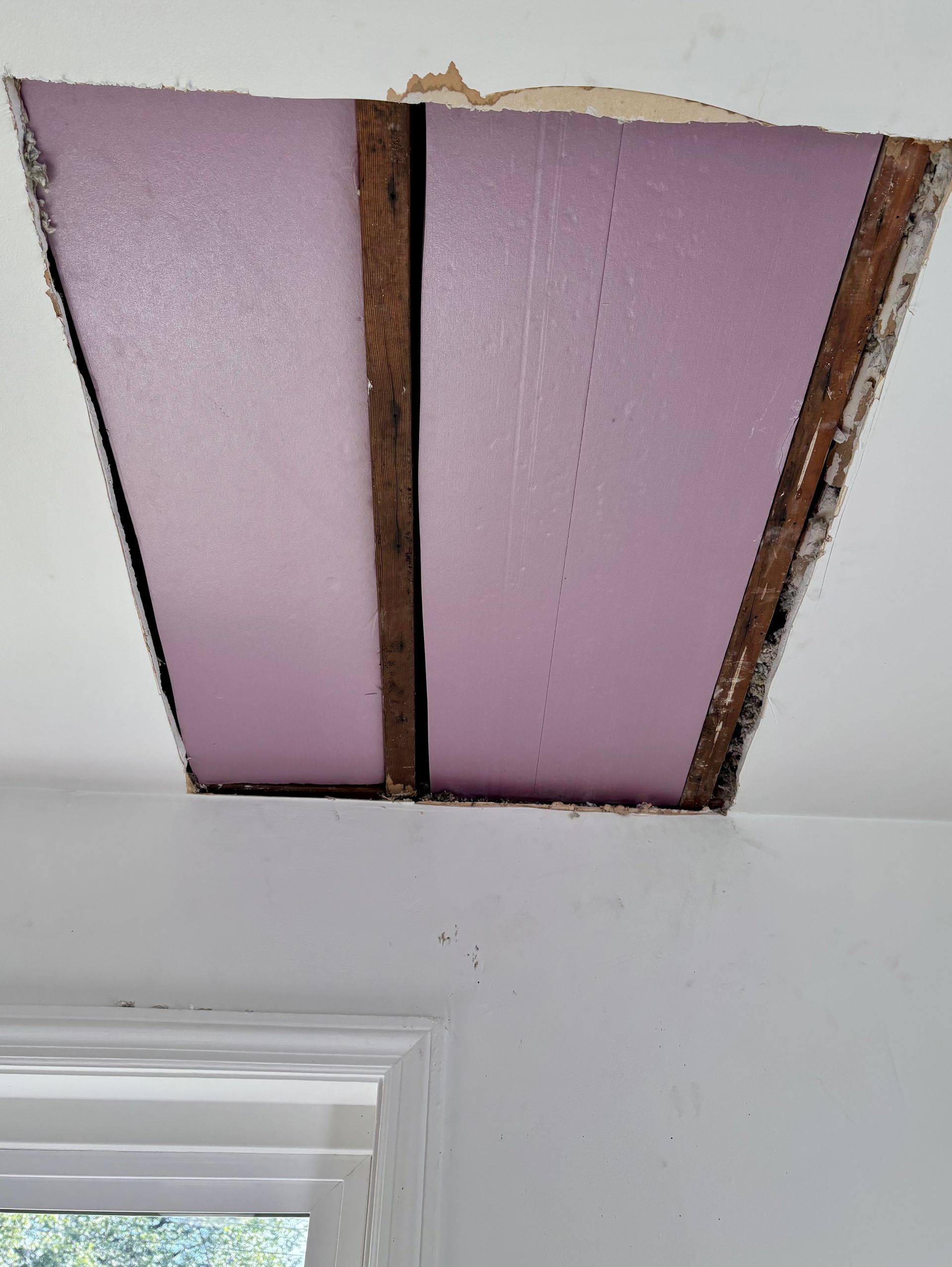 Hole in a white ceiling revealing pink foam insulation and wooden beams.