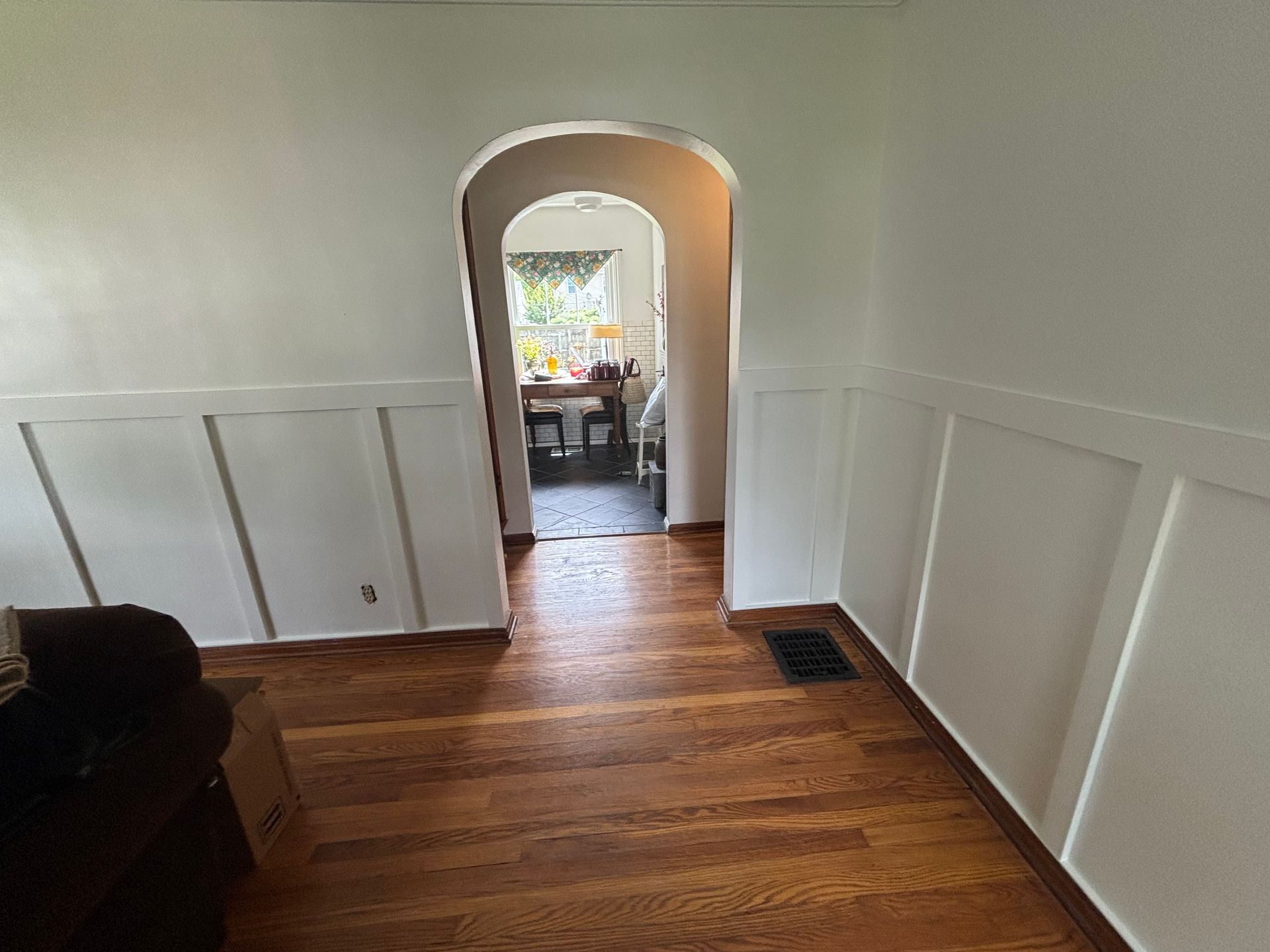 Wooden floor leads to an arched doorway. White wainscoting on walls, looking into another room.