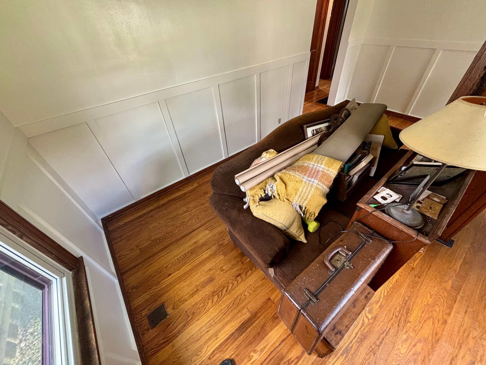 Brown sofa with yellow blanket, lamp, suitcase, and various items in a room with wood floors and white panel walls.