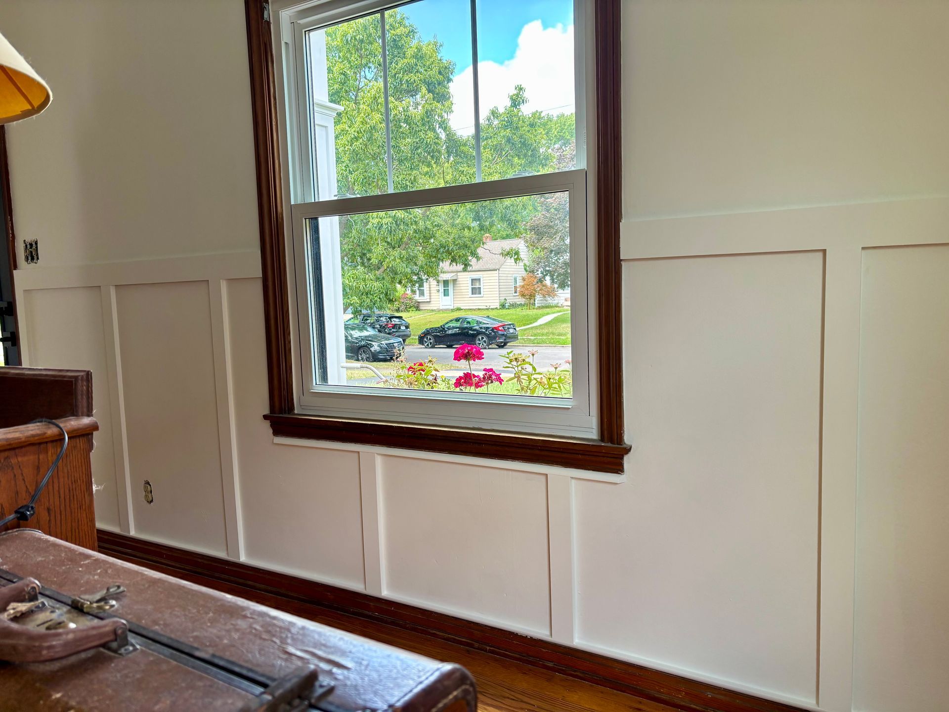 Window with white wall paneling, view of a street with cars, flowers, and trees.