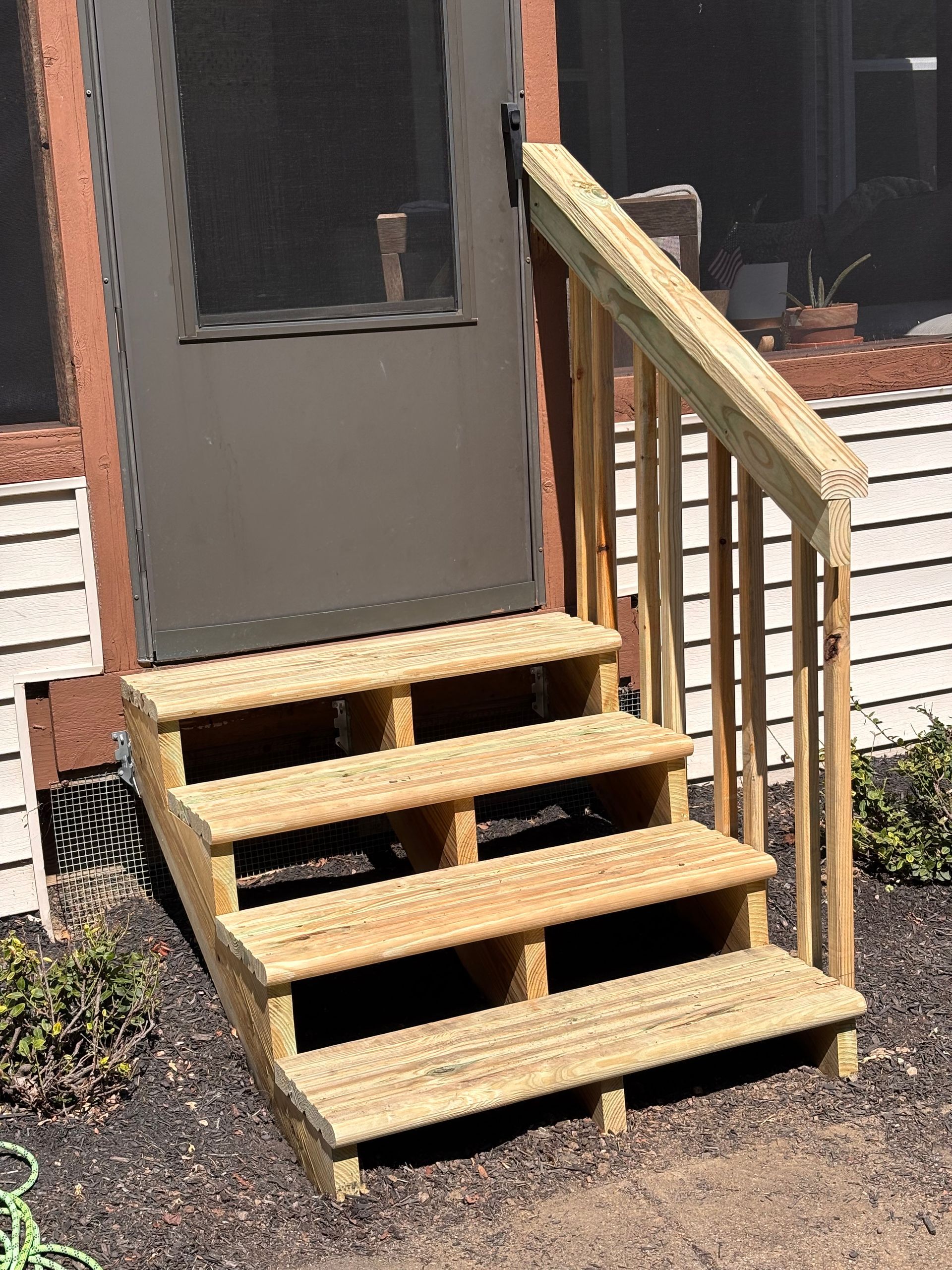 Wooden outdoor steps leading to a dark door, with a handrail on one side.