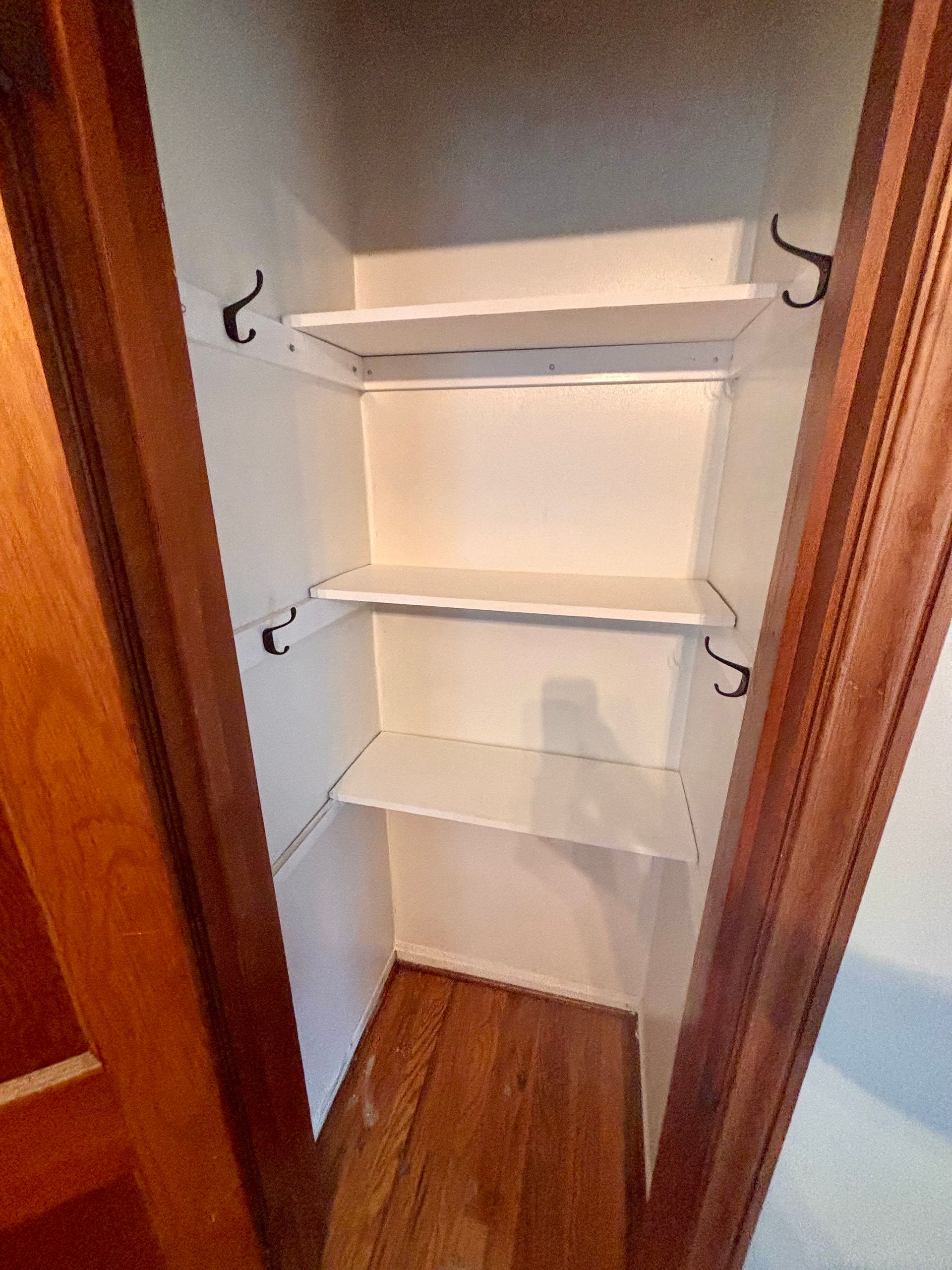 Empty white closet with three shelves and hooks, wooden trim, and hardwood floor.