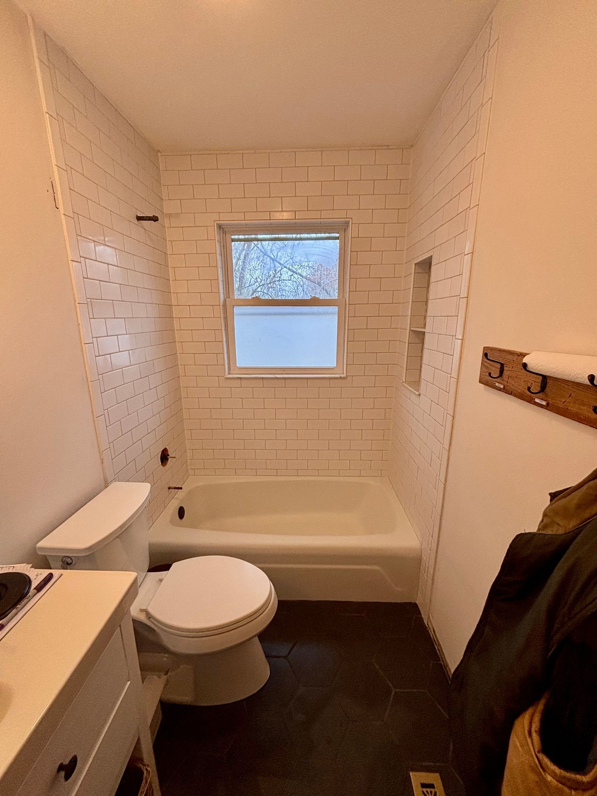 Small bathroom with white tiled walls, bathtub, toilet, and vanity. Dark floor, frosted window.