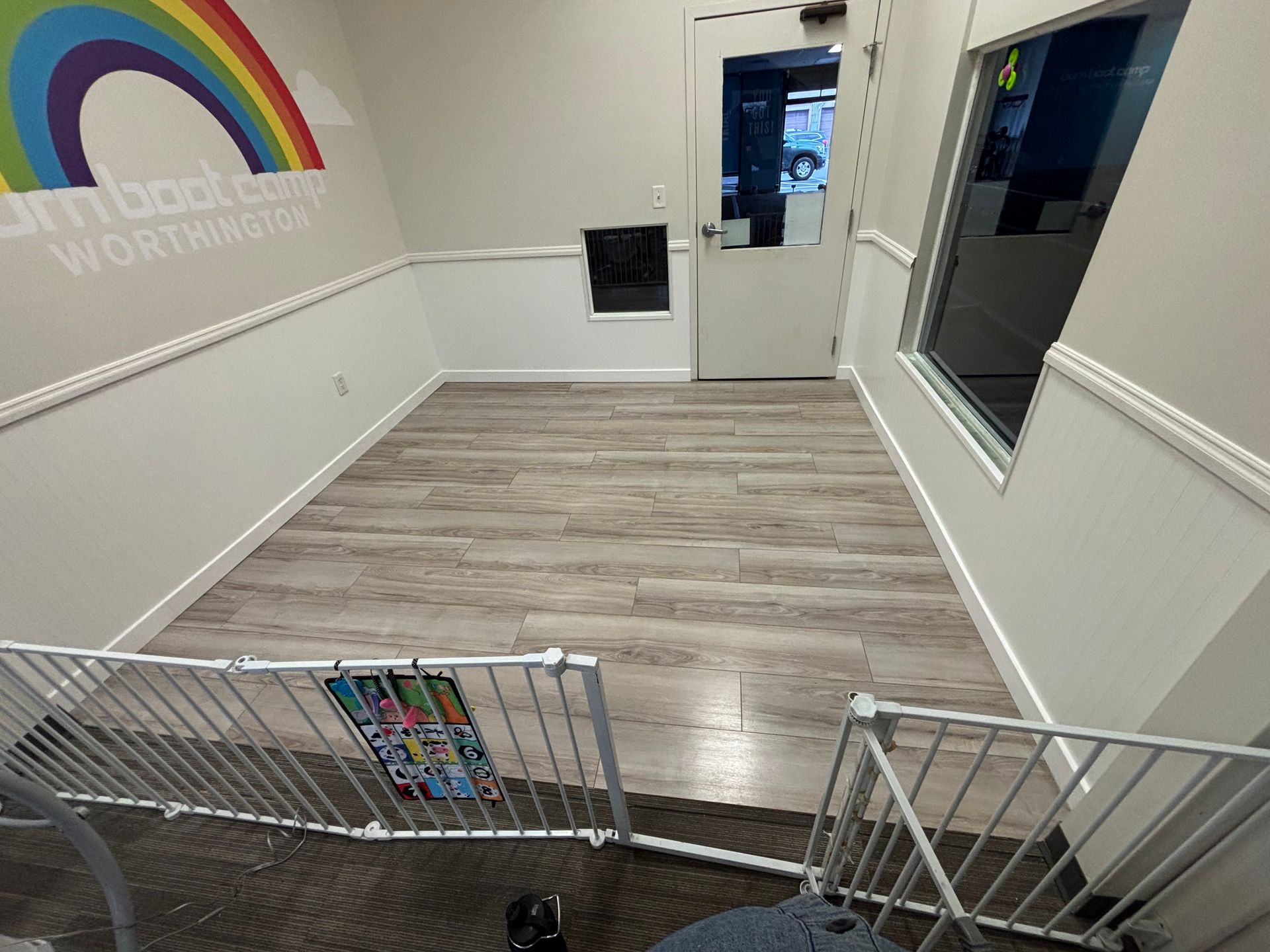 Interior of a room with light wood-look flooring, white walls, and a rainbow logo. A doorway and window are visible.
