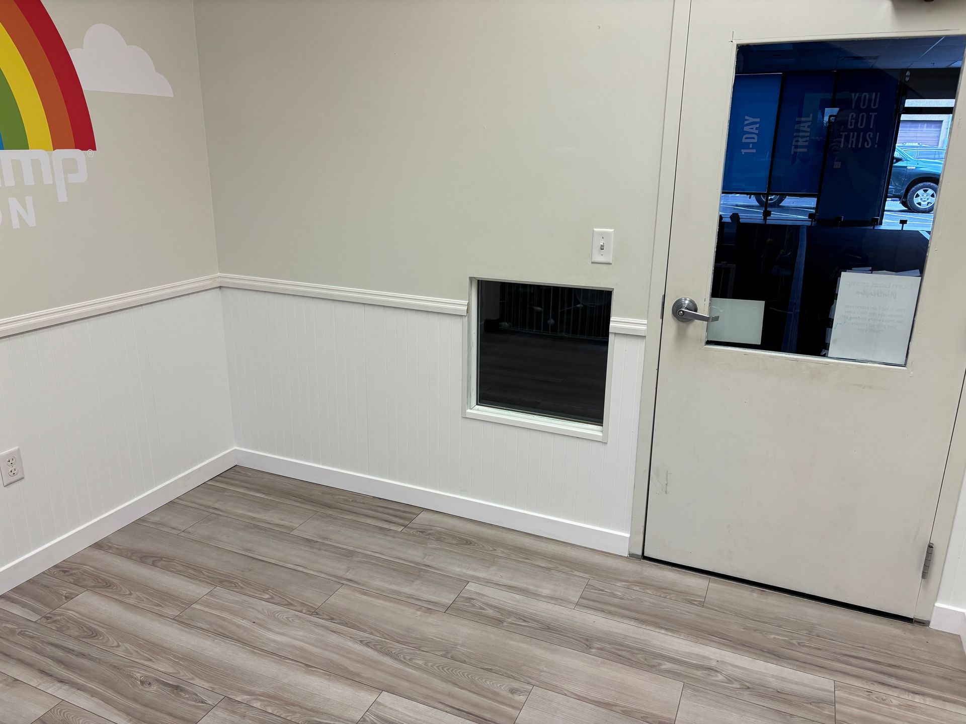 A small room with wood flooring, white wainscoting, a door, and a cutout in the wall. A rainbow is painted on the wall.