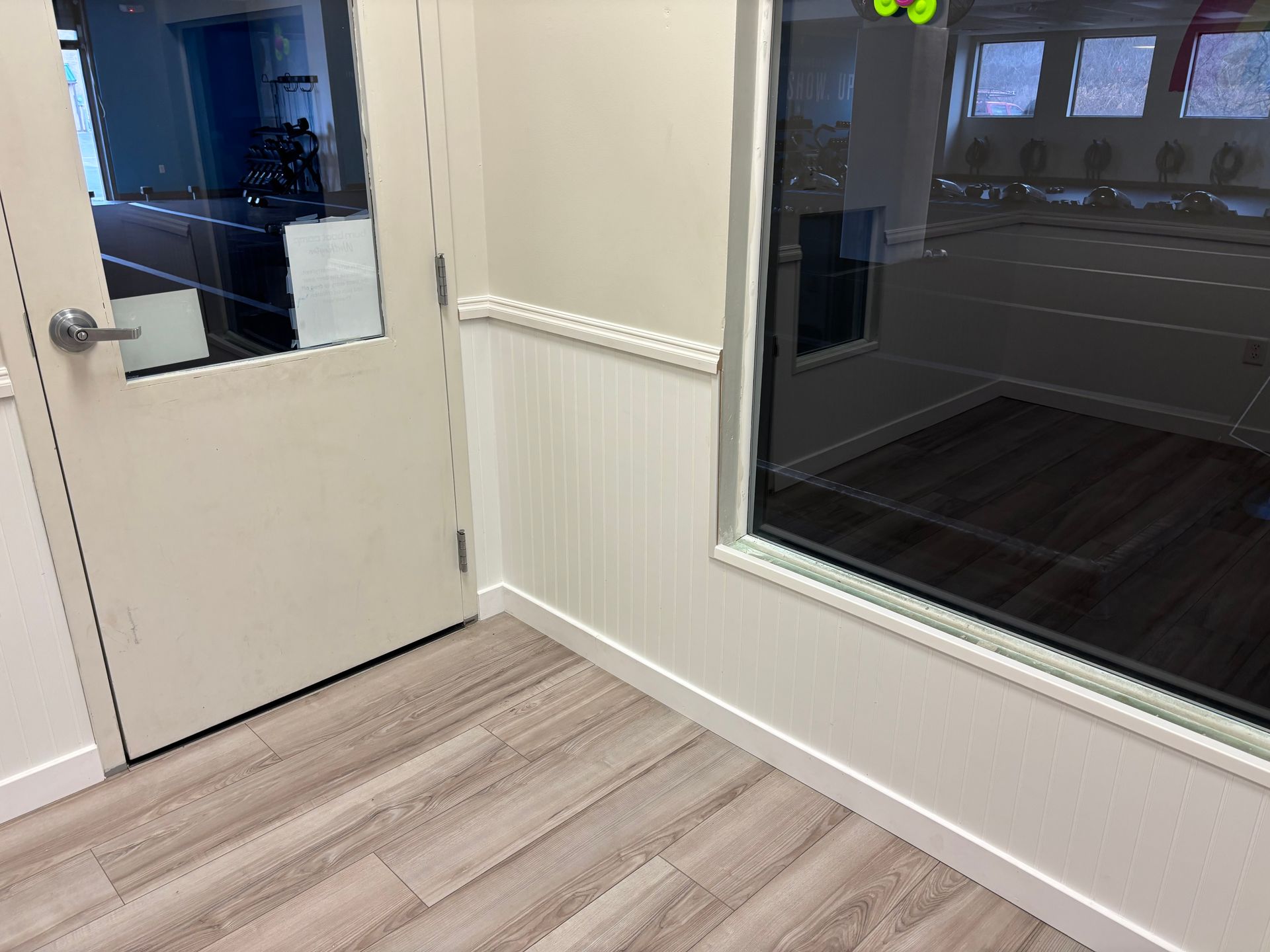 A door and a window in a room with light wood-look flooring. The window has a view of an area with tables.