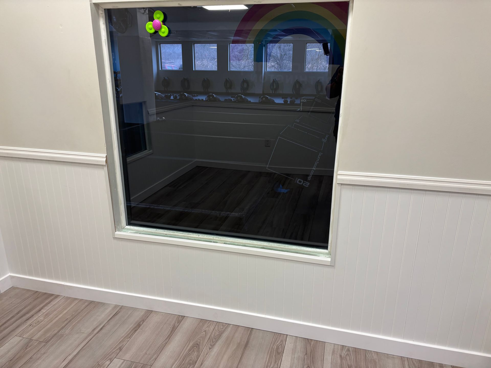 Window looking into a brightly lit room with a rainbow and other decorations; white walls, wood floor.