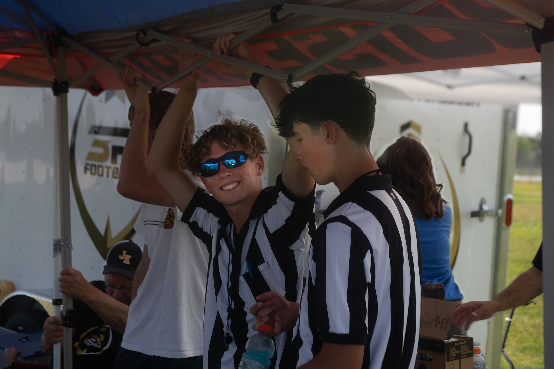 People in black and white striped official shirts stand under a canopy at an outdoor event.