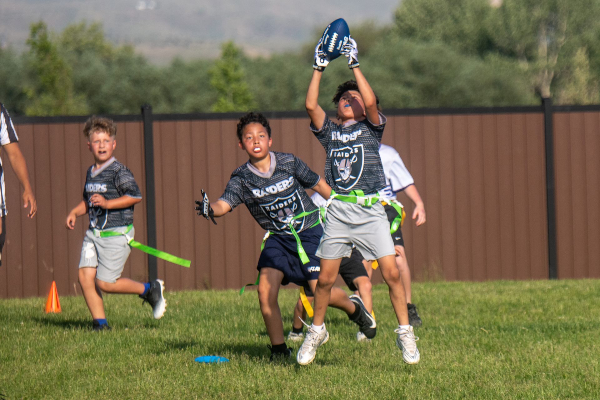Children playing flag football on a grassy field; one player jumps to catch a blue ball while others run nearby.