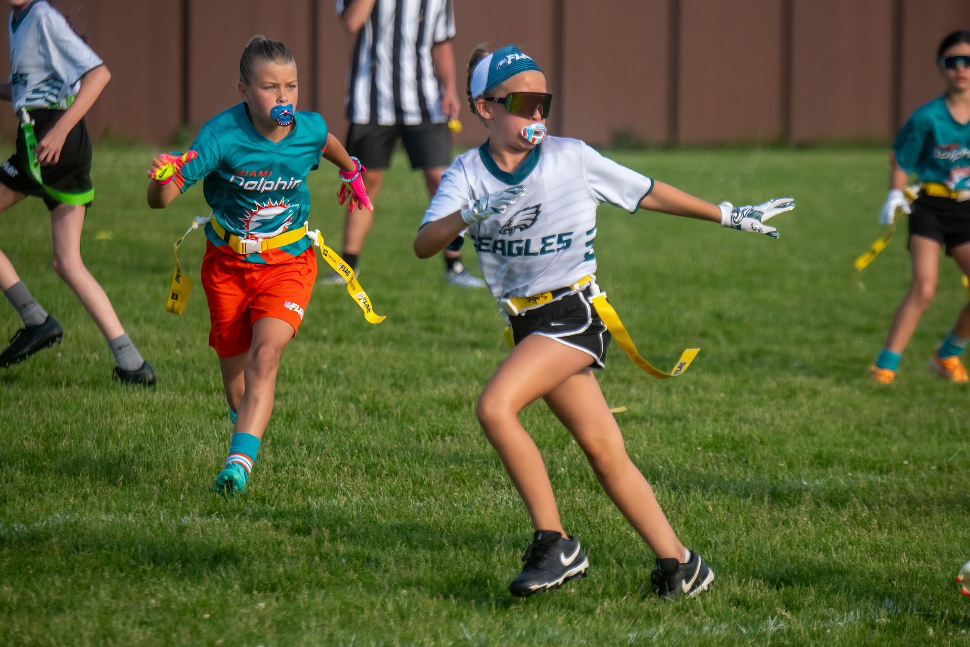 Youth flag football players in teal and white uniforms run on a green field during a game.