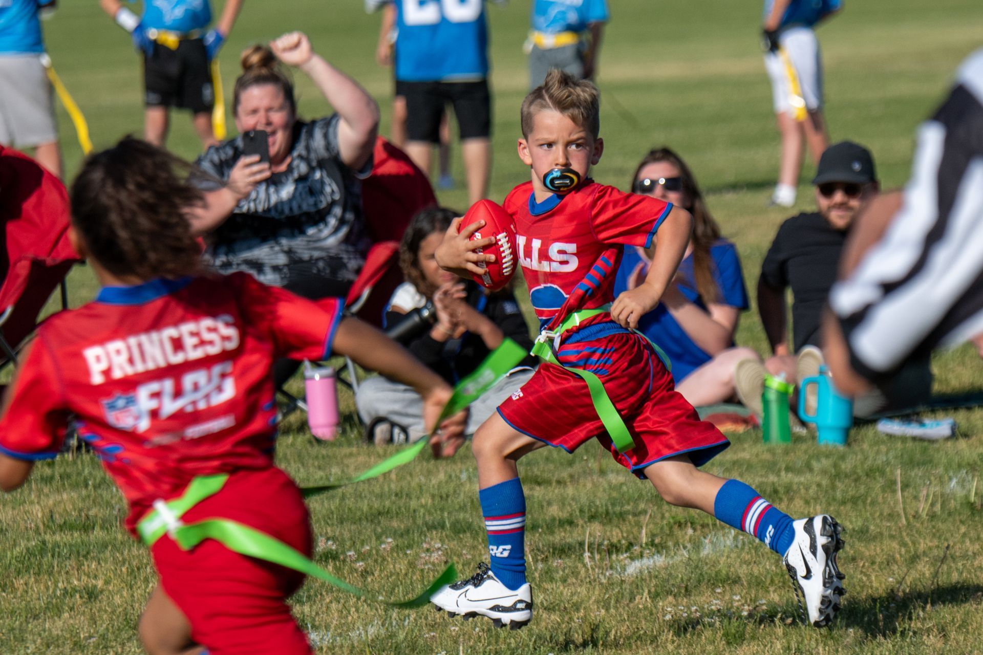 A youth flag football player in a red uniform runs with the ball while another player chases on a sunny grass field.