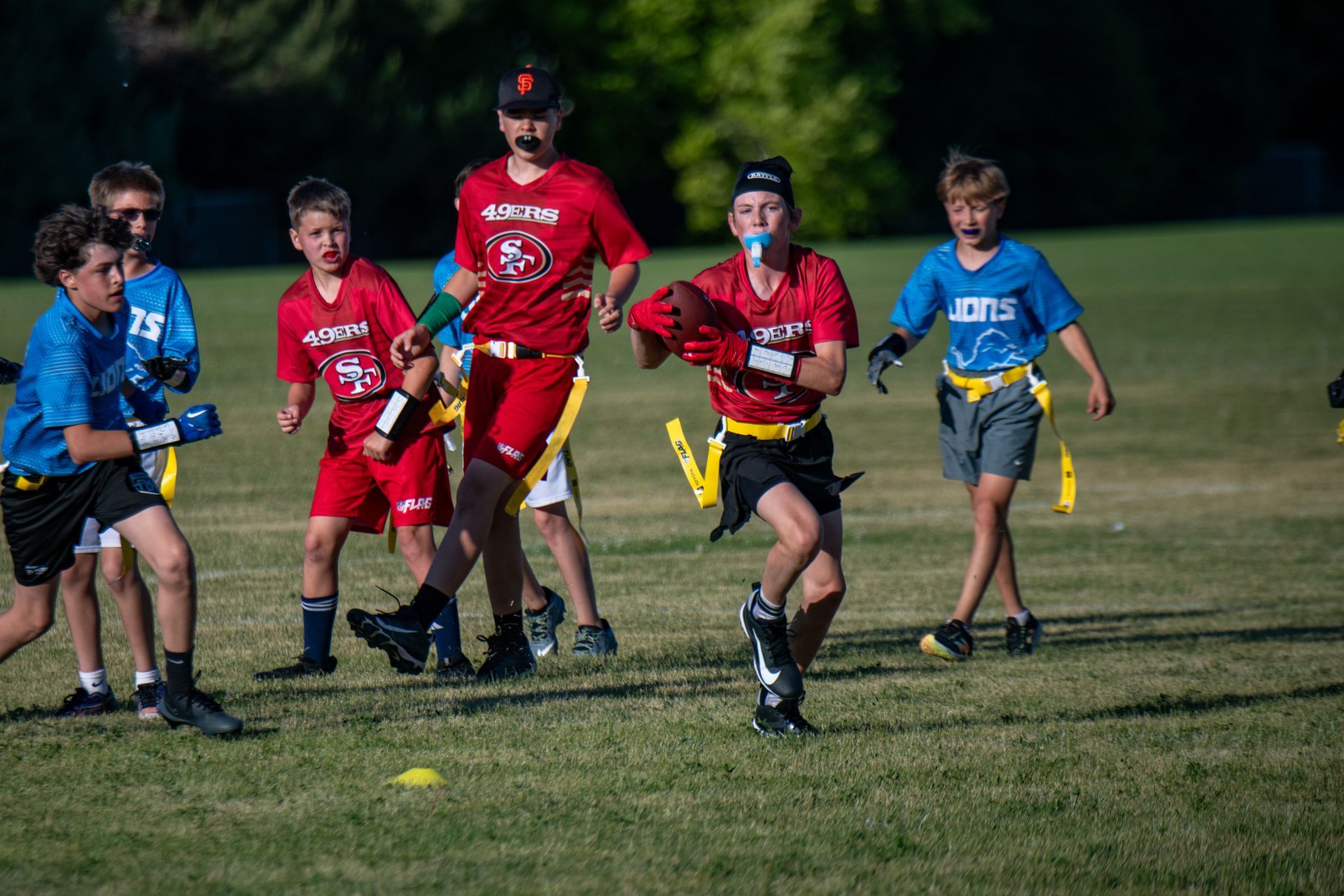 Youth flag football players in red and blue uniforms competing on a grass field during a game.