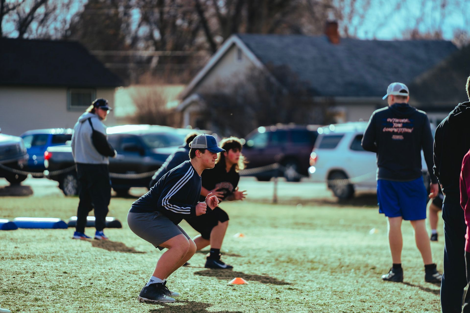 People in athletic wear practice football drills on a grassy field in a suburban setting during the day.