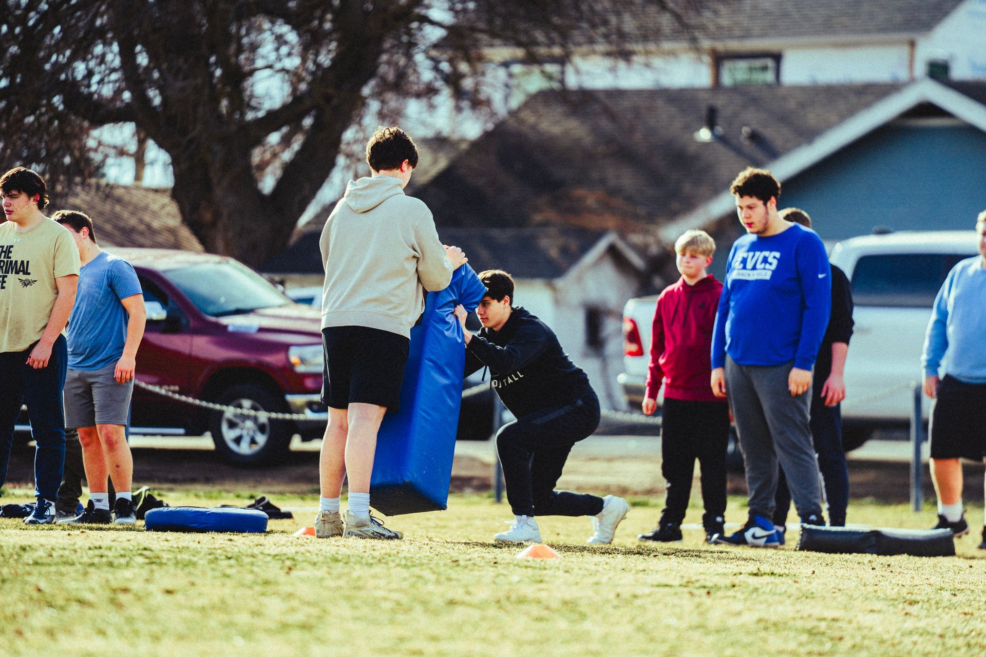A group of students practice football drills with a blue tackling dummy on an outdoor field near parked vehicles.