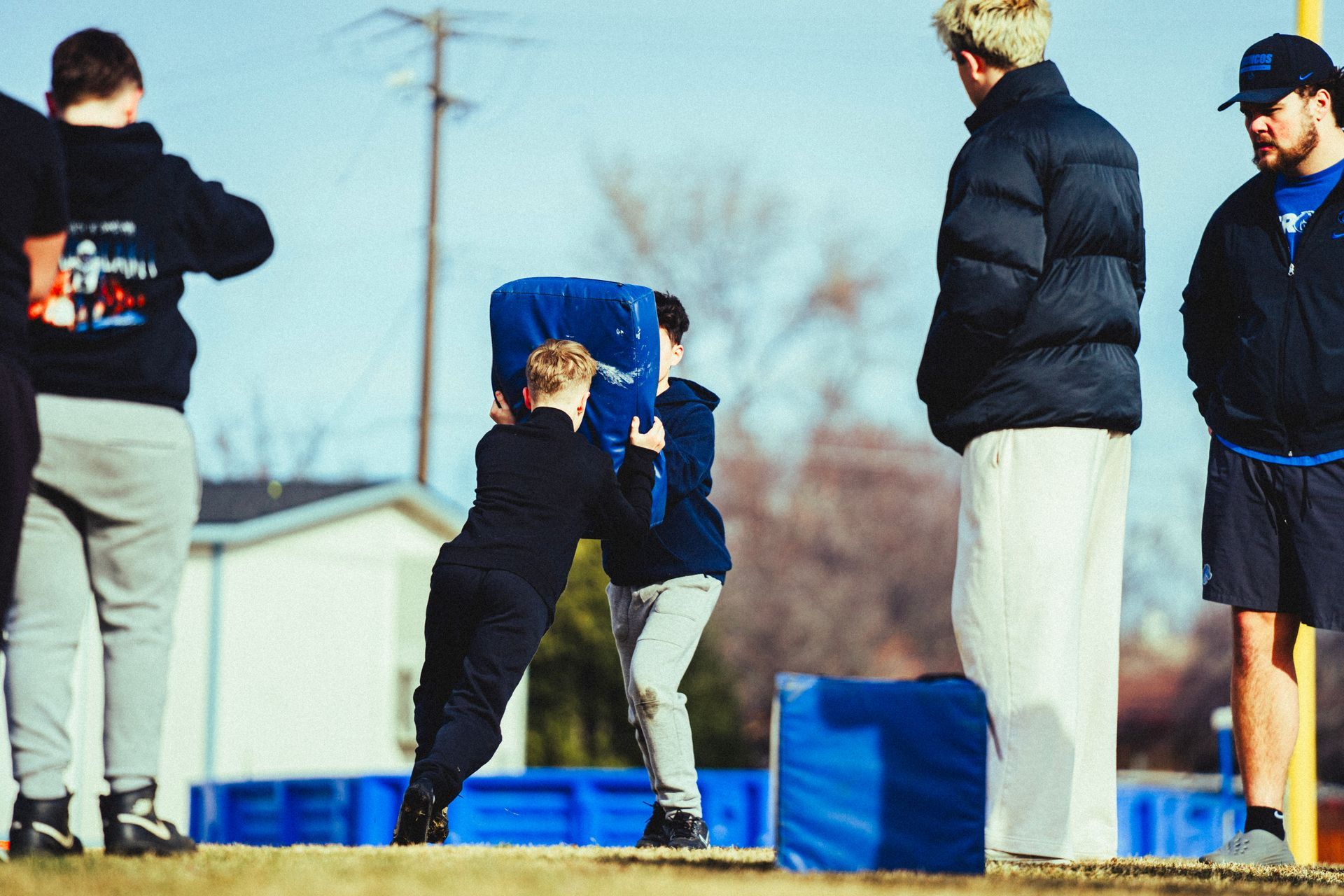 Two people practice football drills using a blue blocking pad on an outdoor field while observers watch nearby.