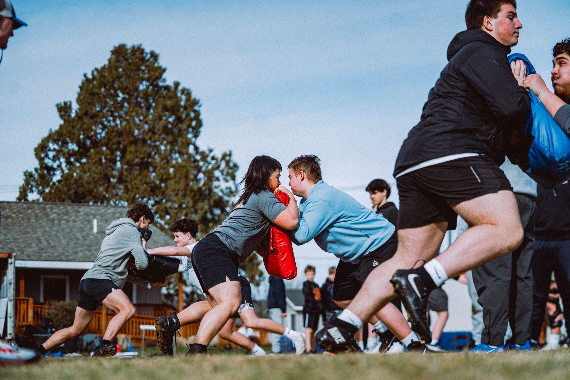 Athletes practice football blocking drills on an outdoor field, using pads to push against each other.