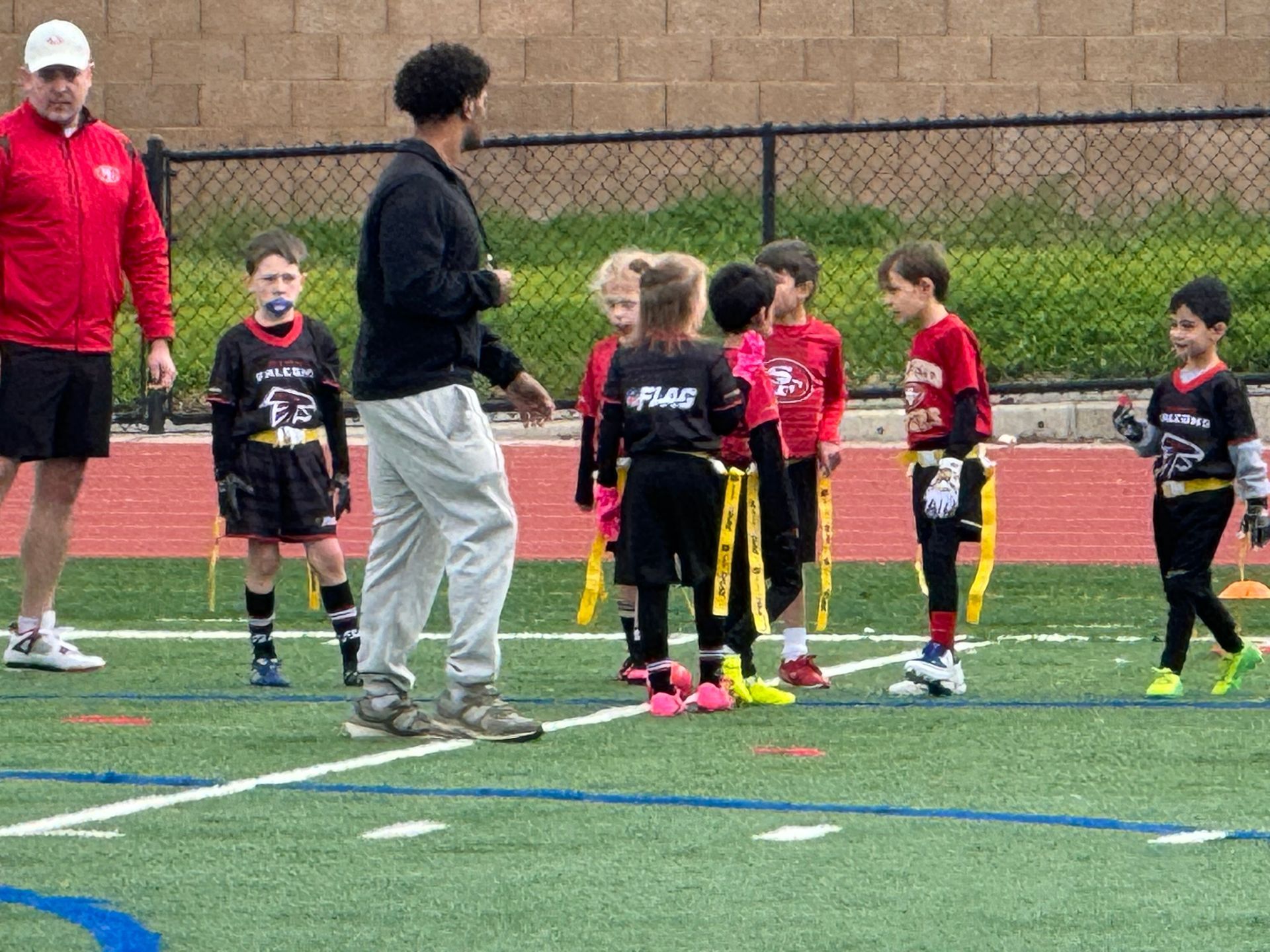 A coach and a group of children in sports uniforms stand on an outdoor field during a flag football practice.