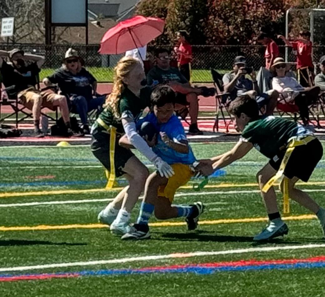 Two flag football players in green jerseys defend against a player in a blue shirt on a field during a game.