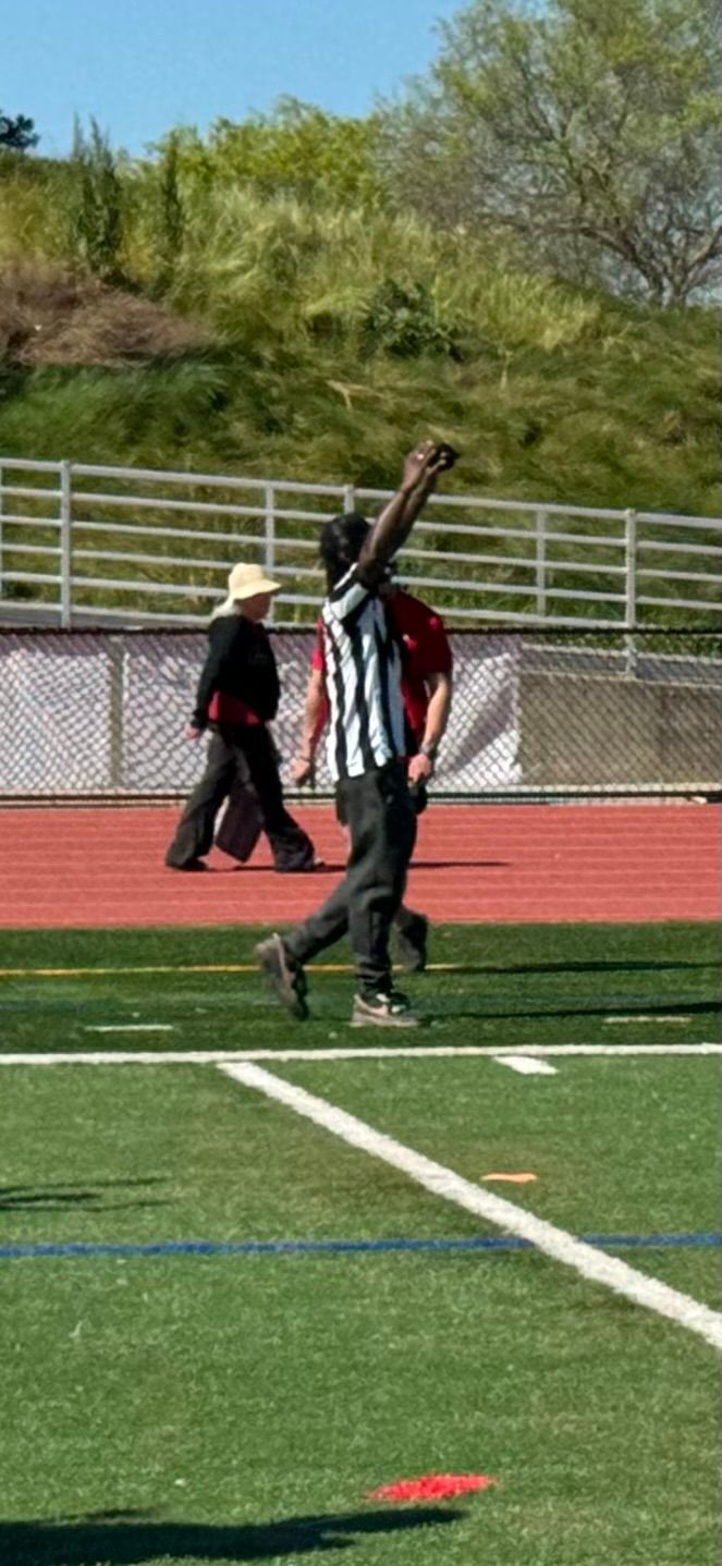 A referee in a striped uniform signals a touchdown on a turf field, with a person in dark clothing walking behind them.