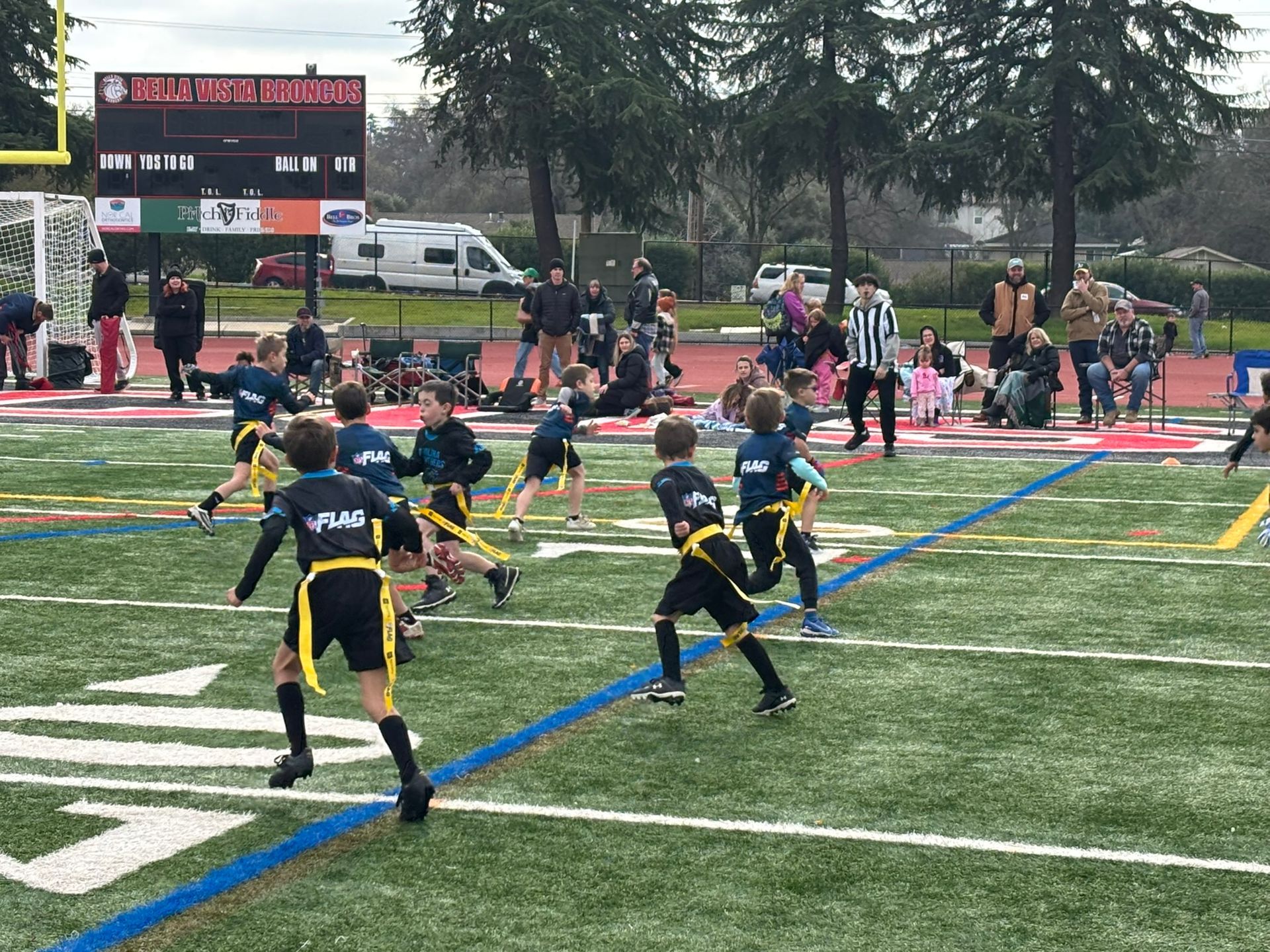 Children in flag football uniforms run across a field during a game on an outdoor turf field with a scoreboard behind them.