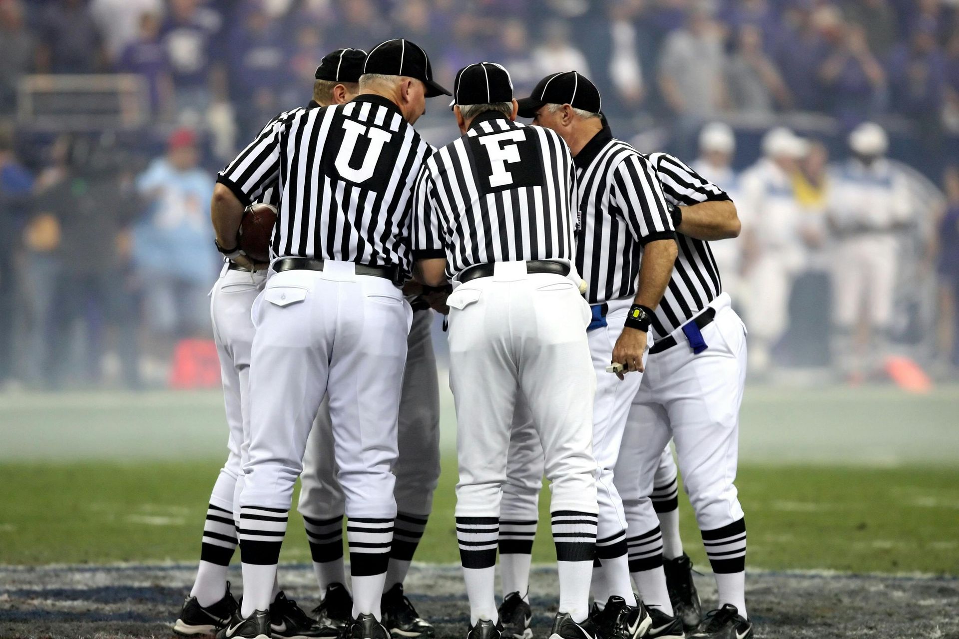 A group of football referees in black-and-white striped shirts stand in a huddle on a grass field during a game.