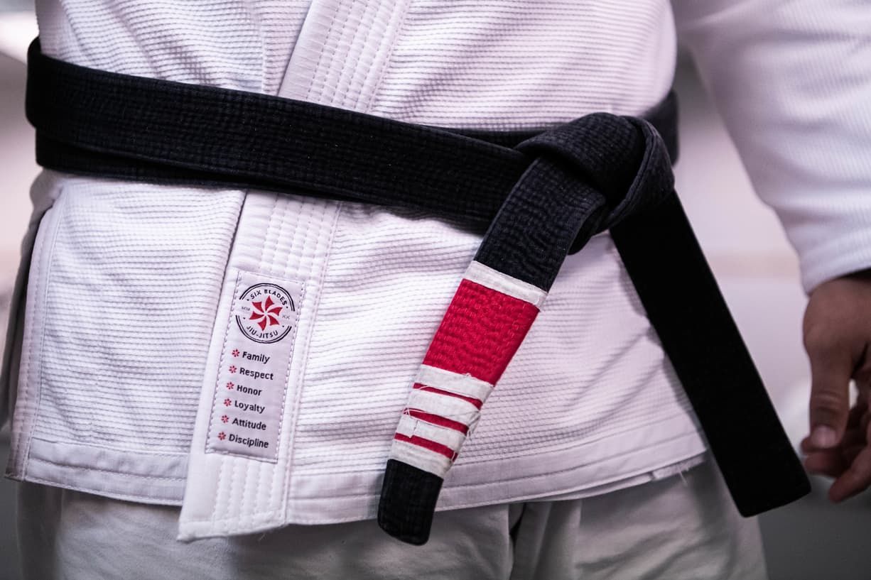 A person wearing a white gi with a black Jiu-Jitsu belt that has a red and white stripe at the end.