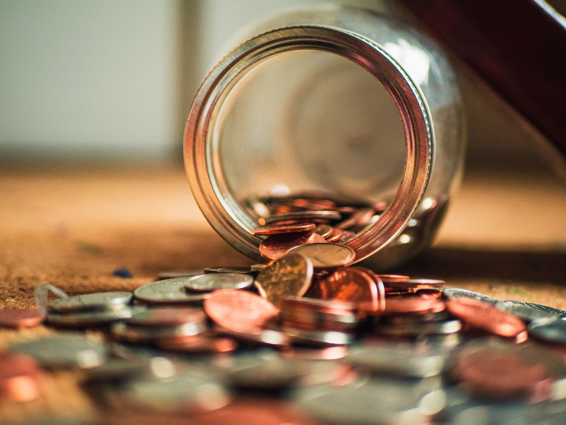 Coins spilling from a clear glass jar onto a wooden surface. Pennies, nickels, and dimes are visible.