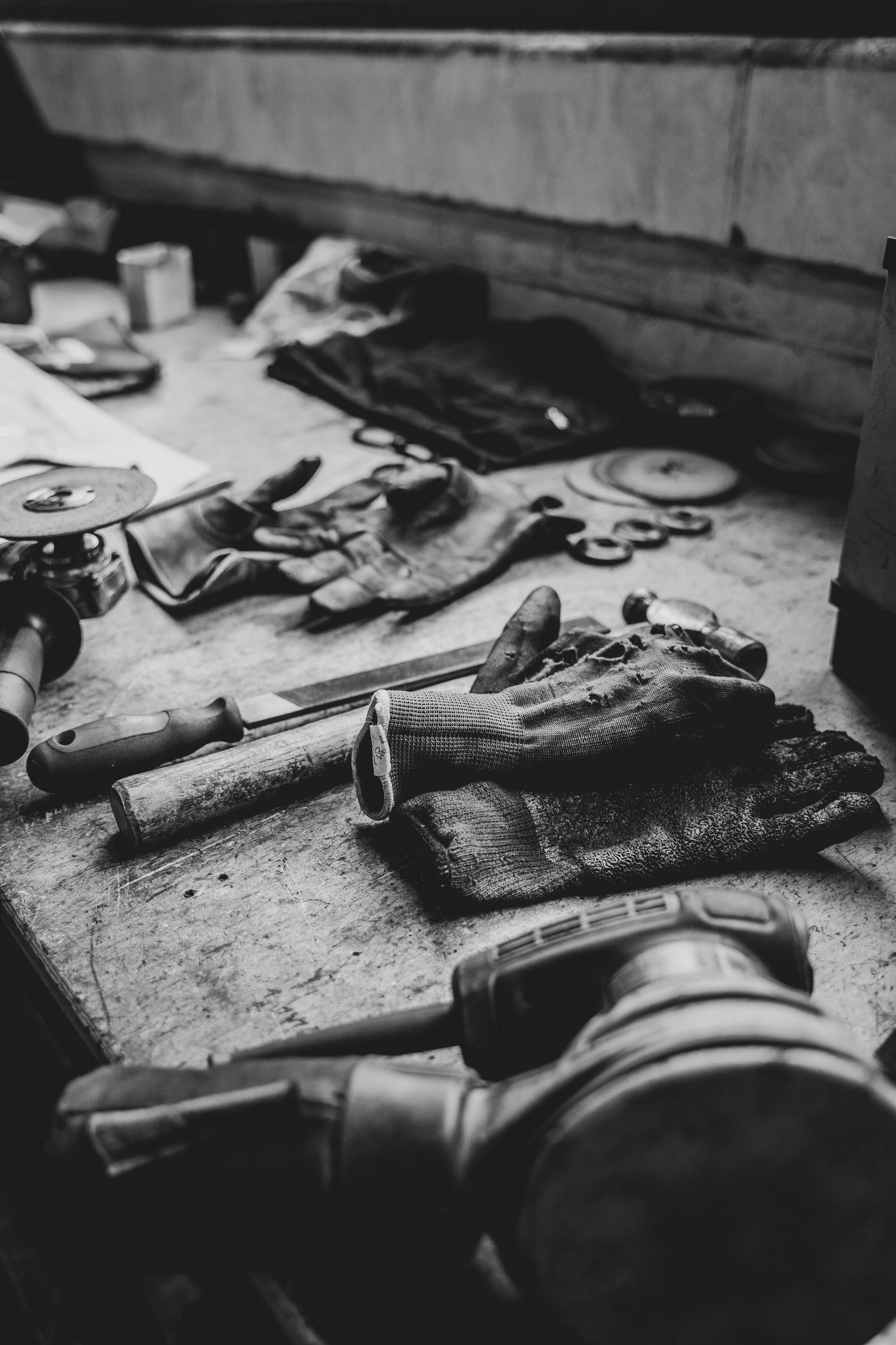 Black and white close-up of a cluttered workbench with tools, gloves, and grinding discs, suggesting a workshop environment.