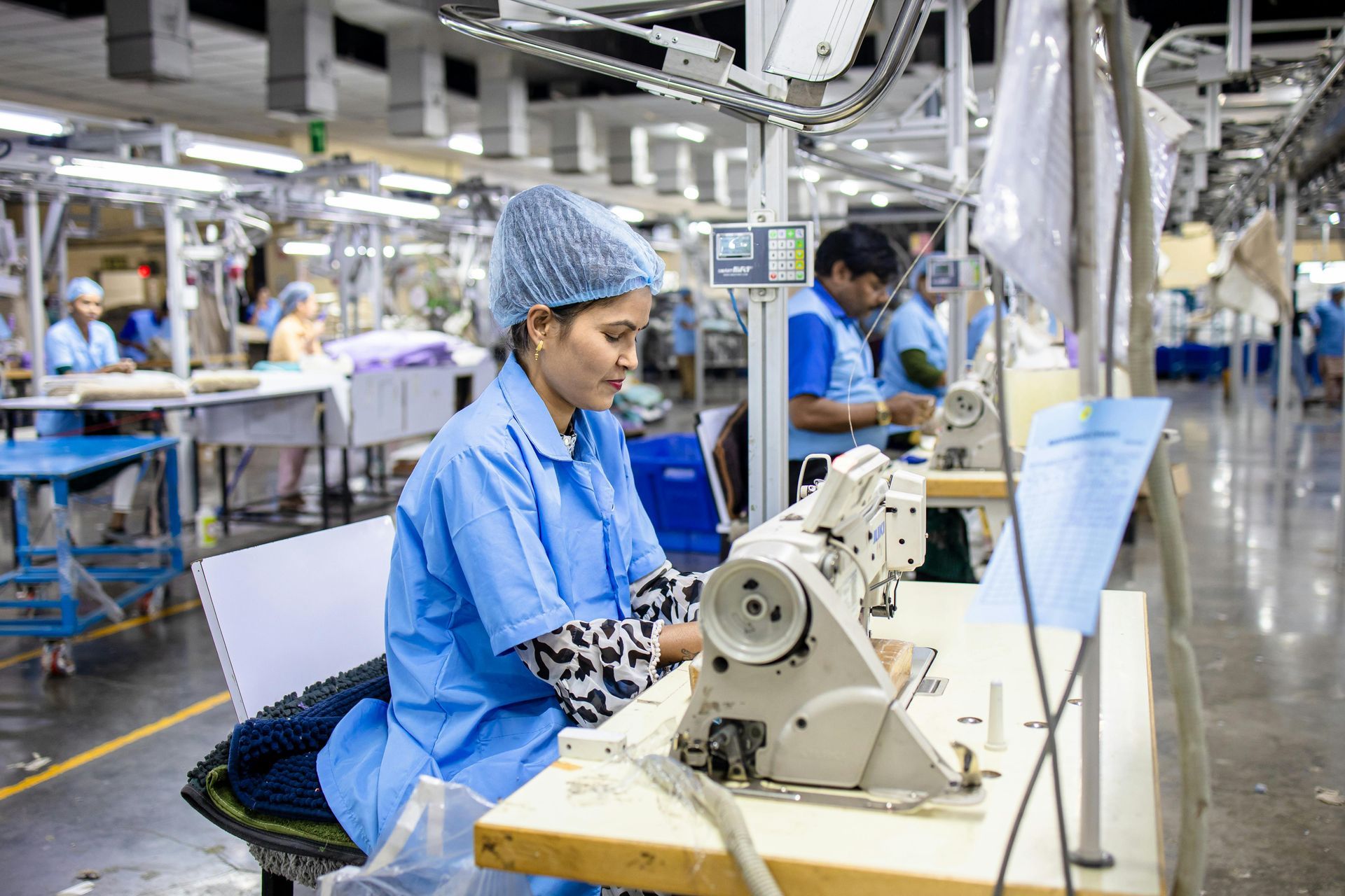 Woman in blue scrubs sews fabric at a factory workstation. Other workers are in the background, all focused on their tasks in a well-lit space.