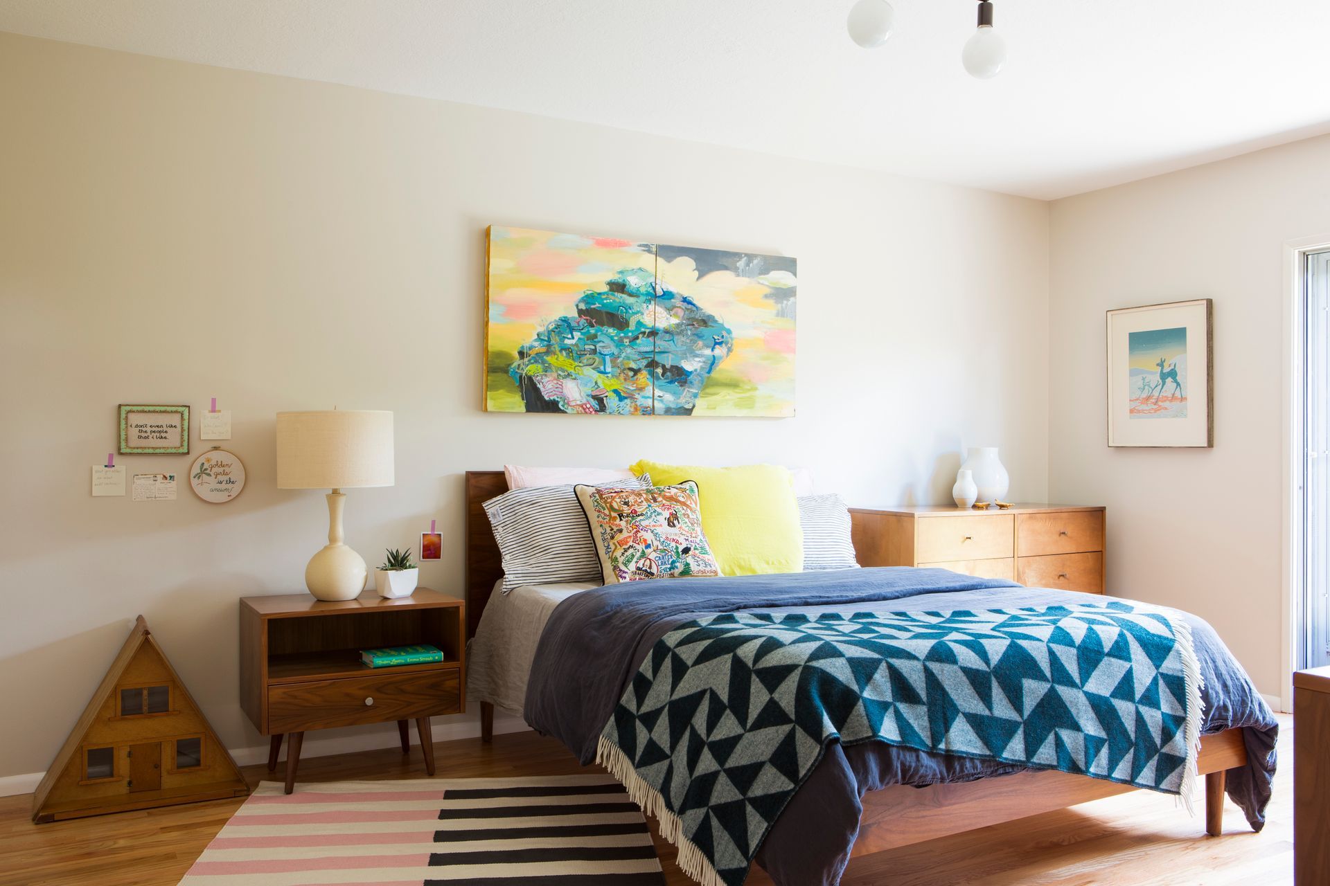 Light-filled bedroom with layered bedding, wood furniture, and colorful artwork in a calm modern interior