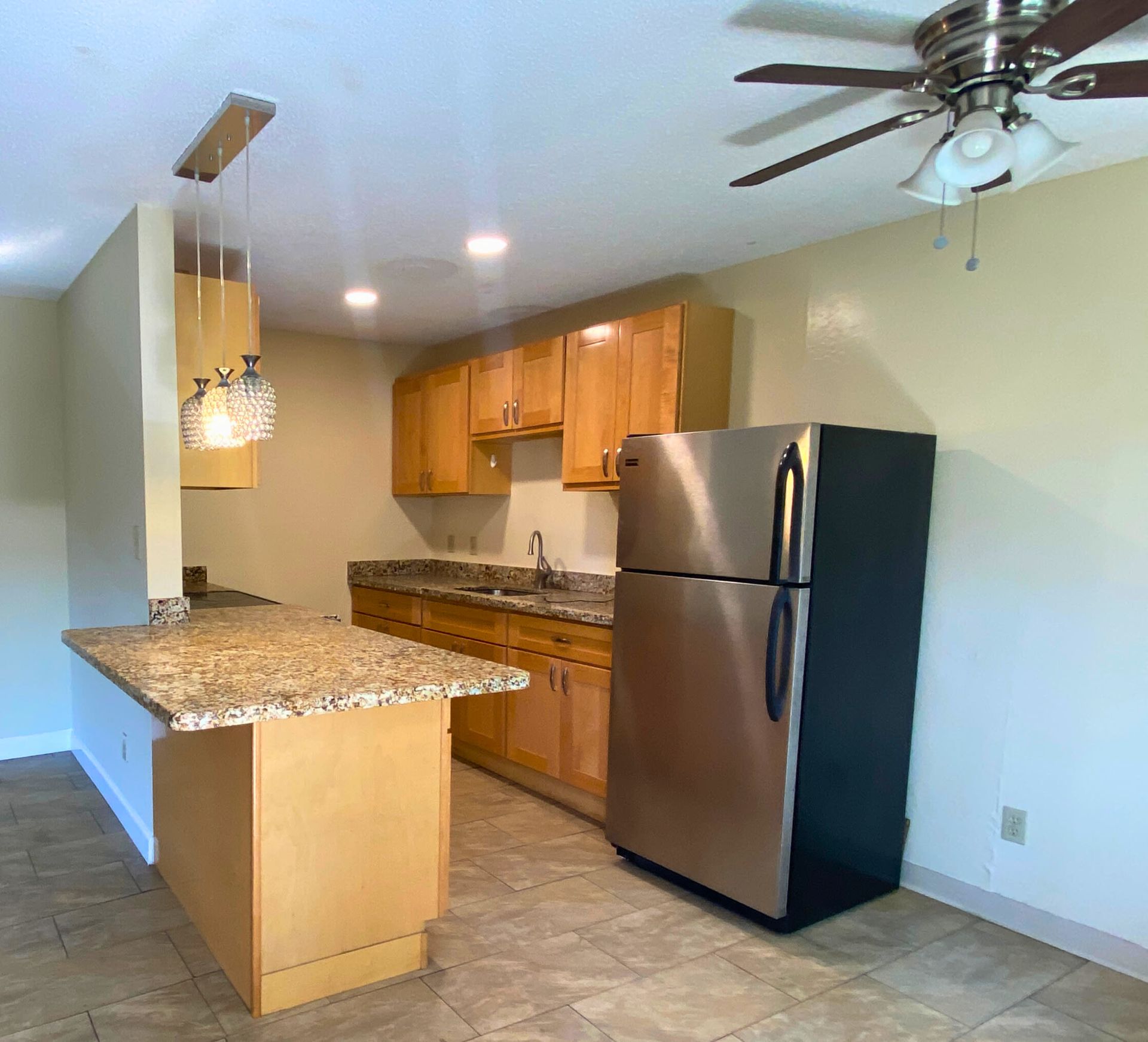 Apartment kitchen with granite countertops, wooden cabinets, stainless refrigerator, island, and ceiling fan.