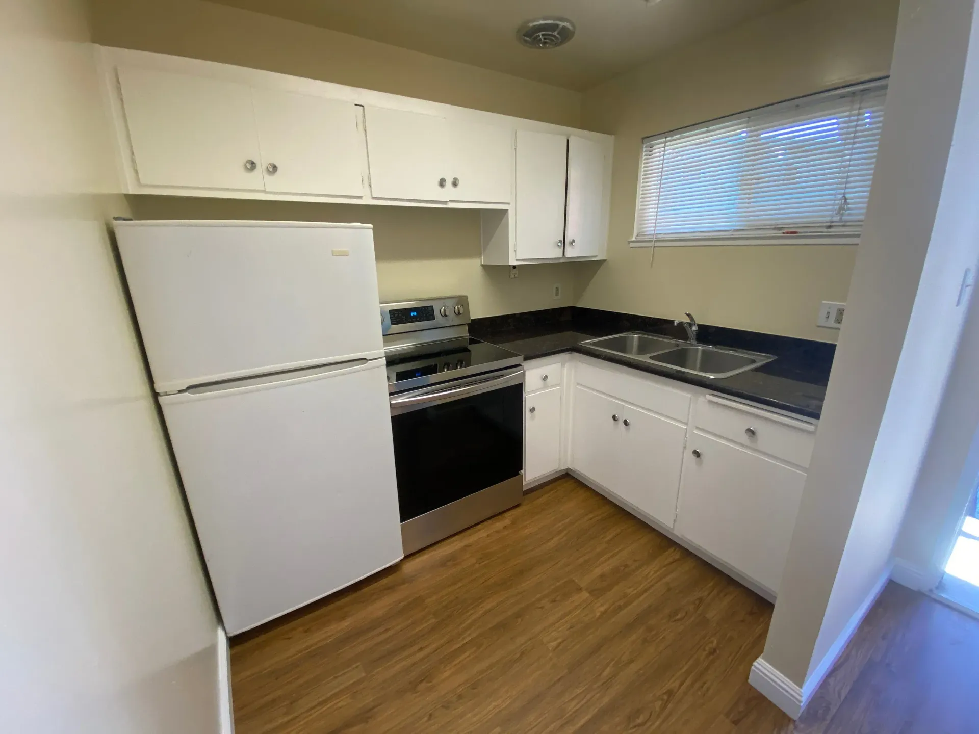 L-shaped kitchen with white cabinets, fridge, stove, double sink, and window with blinds.