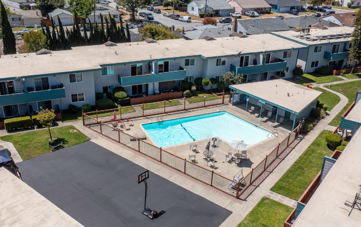 Aerial view of an apartment pool area with lounge chairs, a fence, and blue balconies.