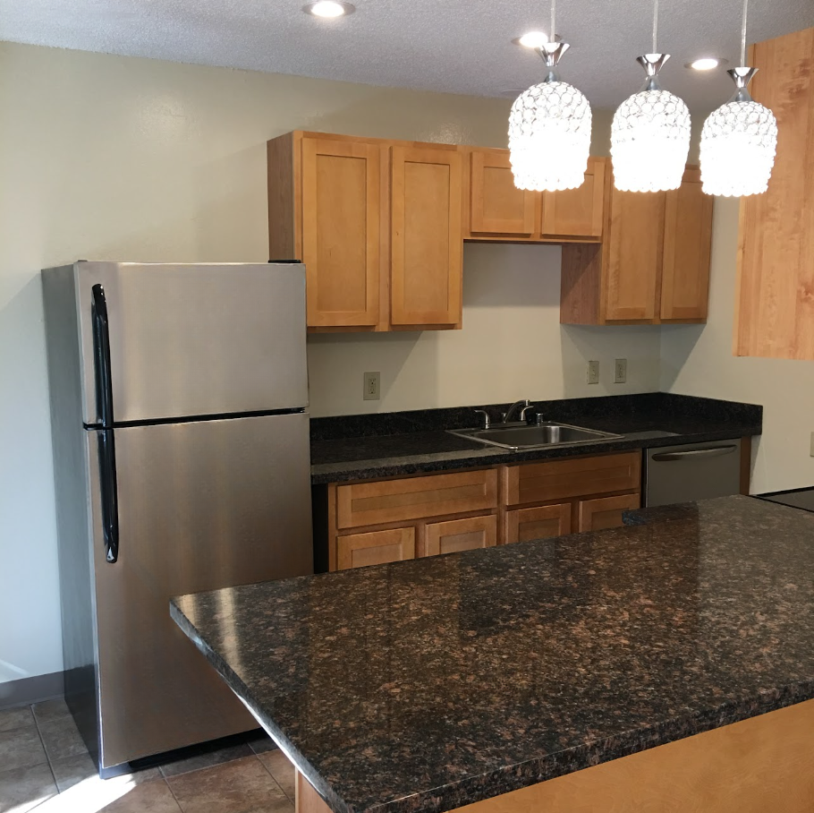 Apartment kitchen with stainless steel refrigerator, wooden cabinets, granite countertops, and an island.