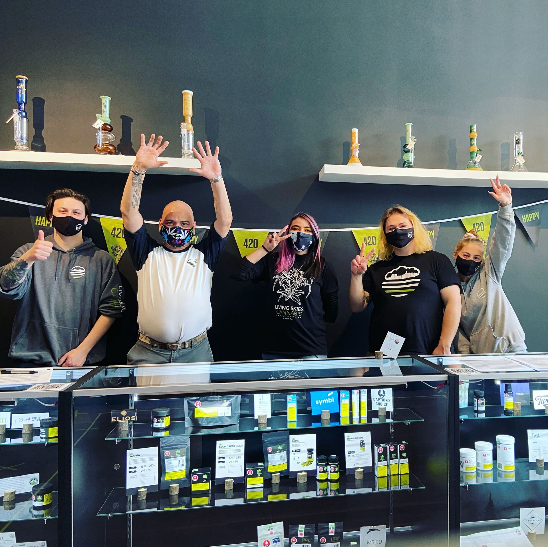 People behind a store counter with products in the display case, waving and wearing masks. Bongs on the shelf.