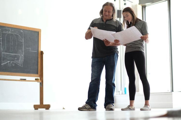 Two people standing by a chalkboard, reviewing large papers in a bright room.
