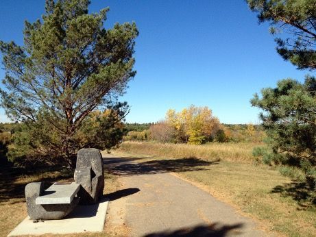 A stone bench sits beside a paved path winding through a grassy field under a clear blue sky, surrounded by pine trees. Saskatoon.